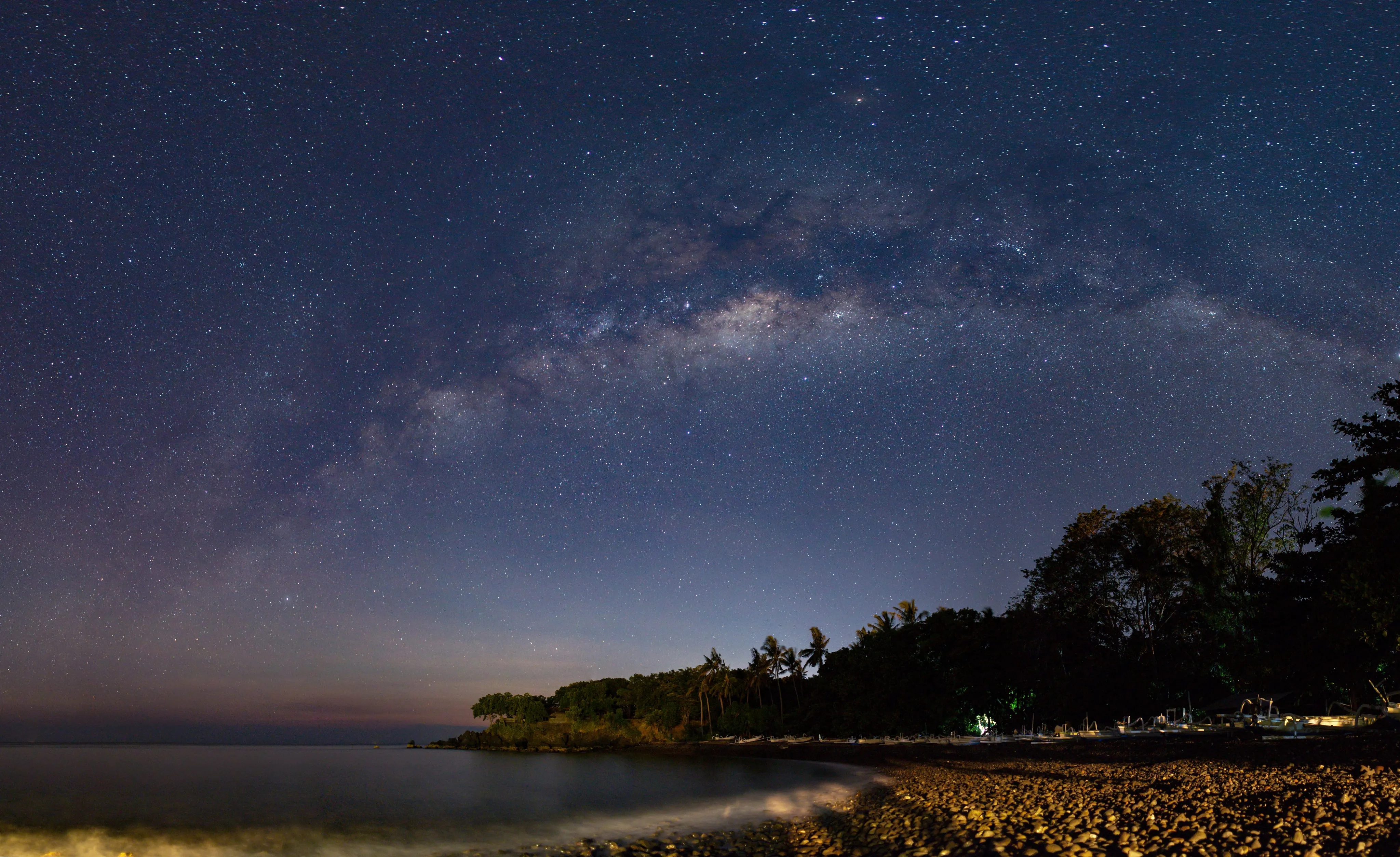 The Milky Way arching over a pebbled beach and wooded shoreline