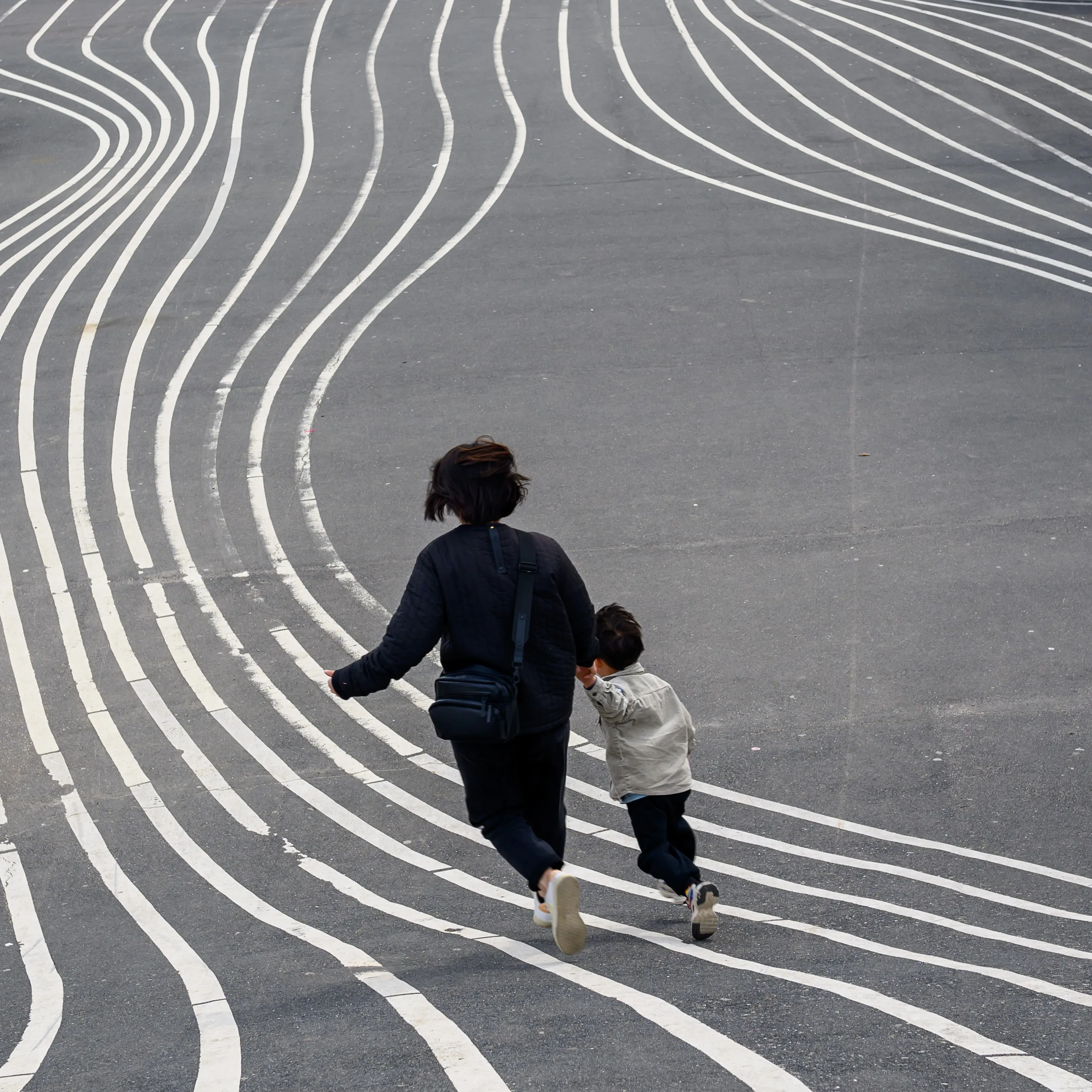 An adult and a child running hand in hand across a plaza painted with curving white lines