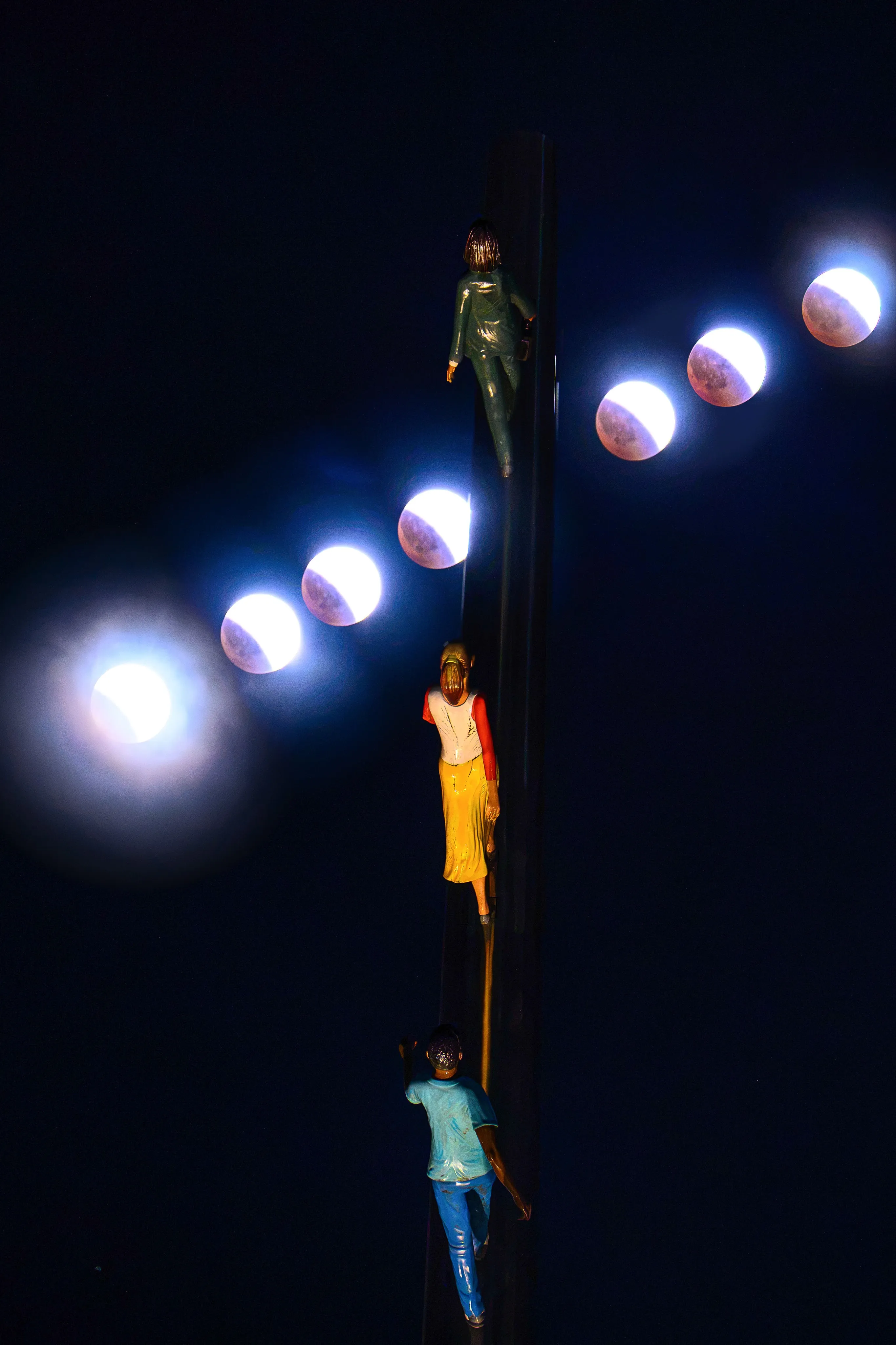 A composite of a lunar eclipse's progression arcing beside the painted climbing figures of the "Walking to the Sky" sculpture.