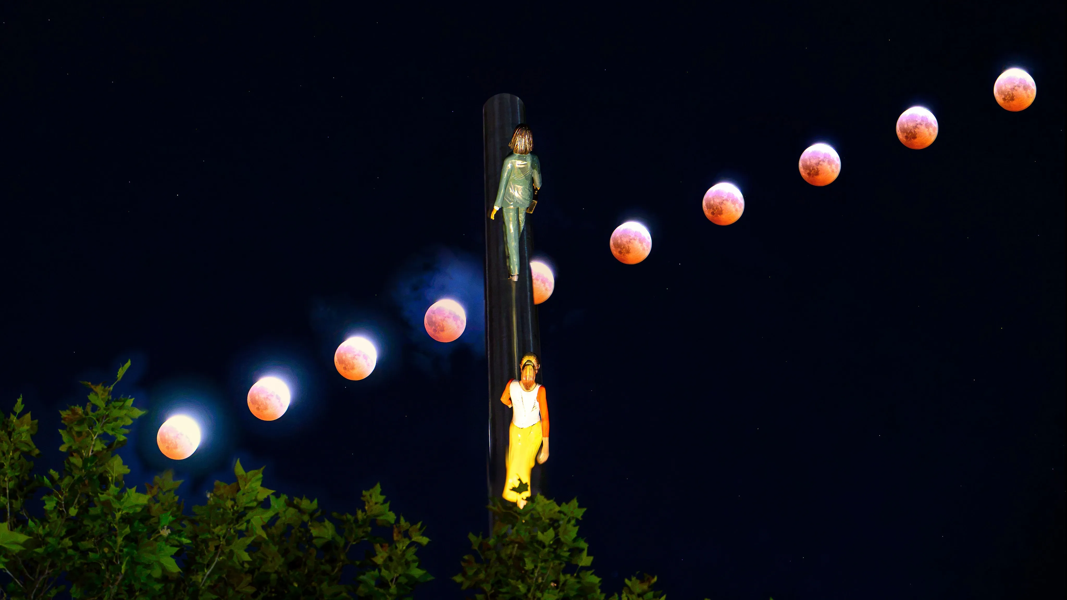 A composite of a blood moon eclipse's progression arcing above treetops beside the "Walking to the Sky" sculpture.