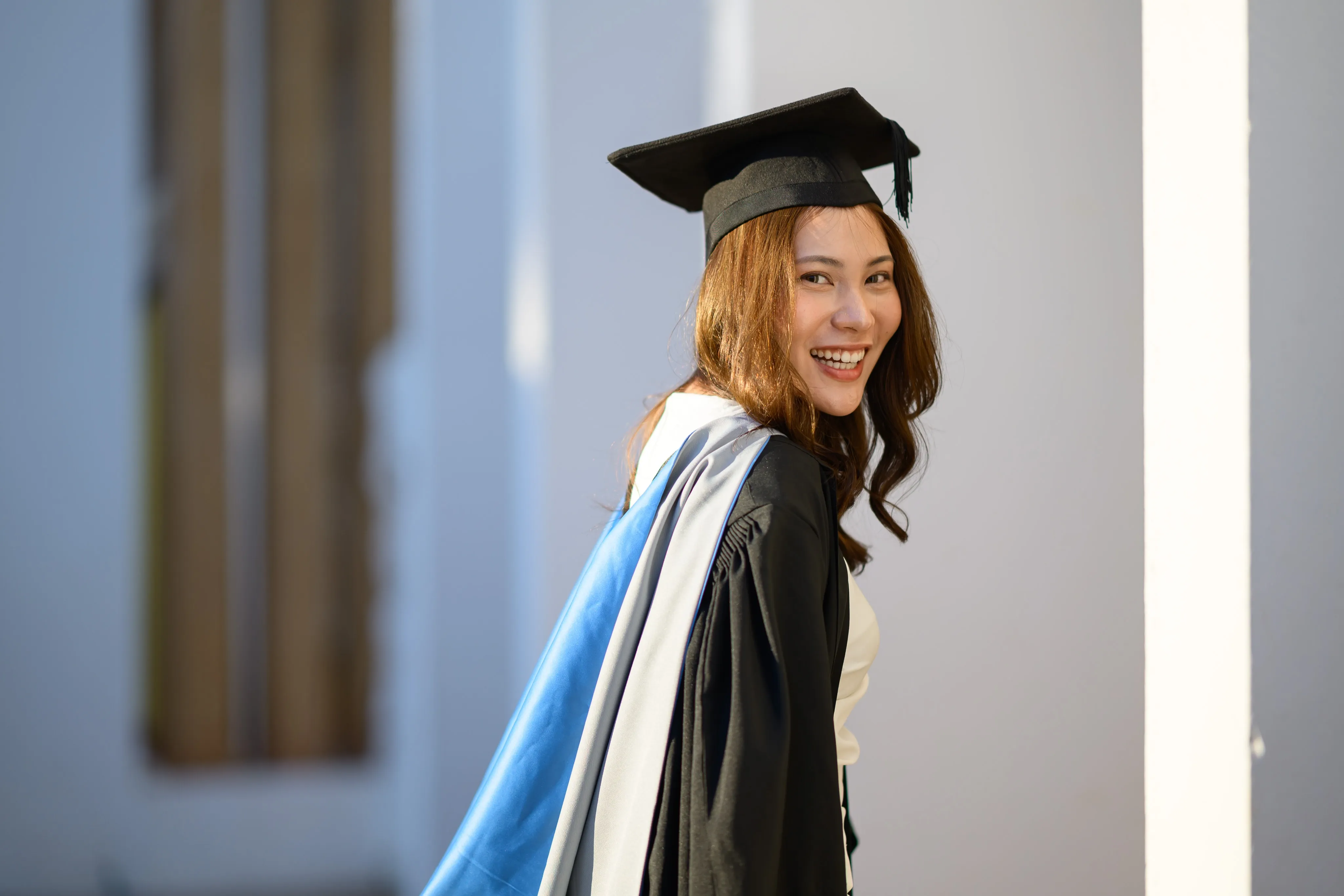 A smiling graduate in cap and gown looking back over her shoulder beside a white building