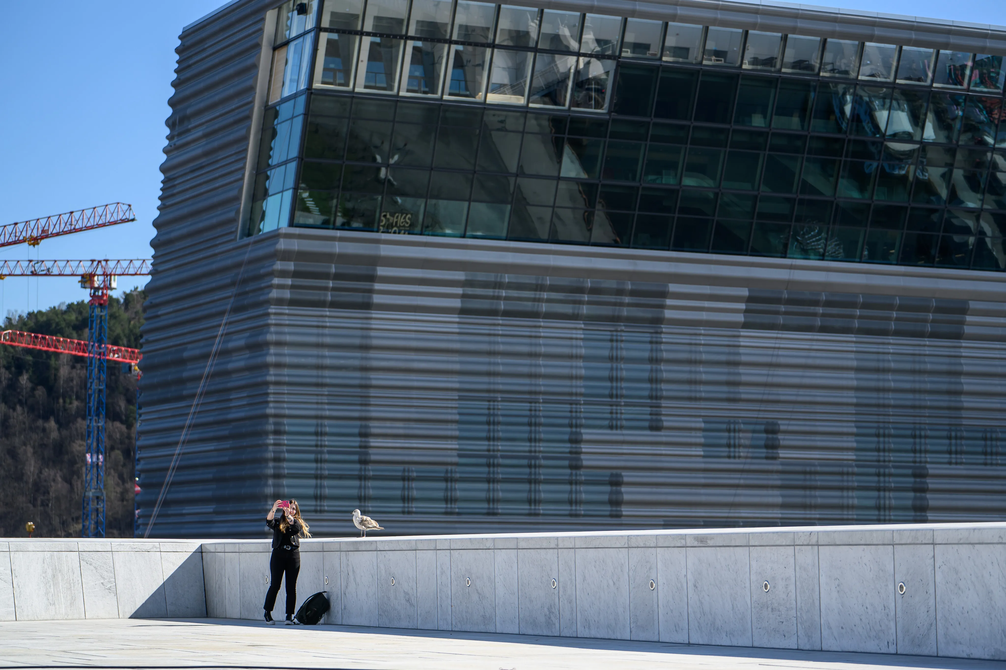 A person taking a photo on a pale stone plaza beneath a striped modern building, a construction crane beyond