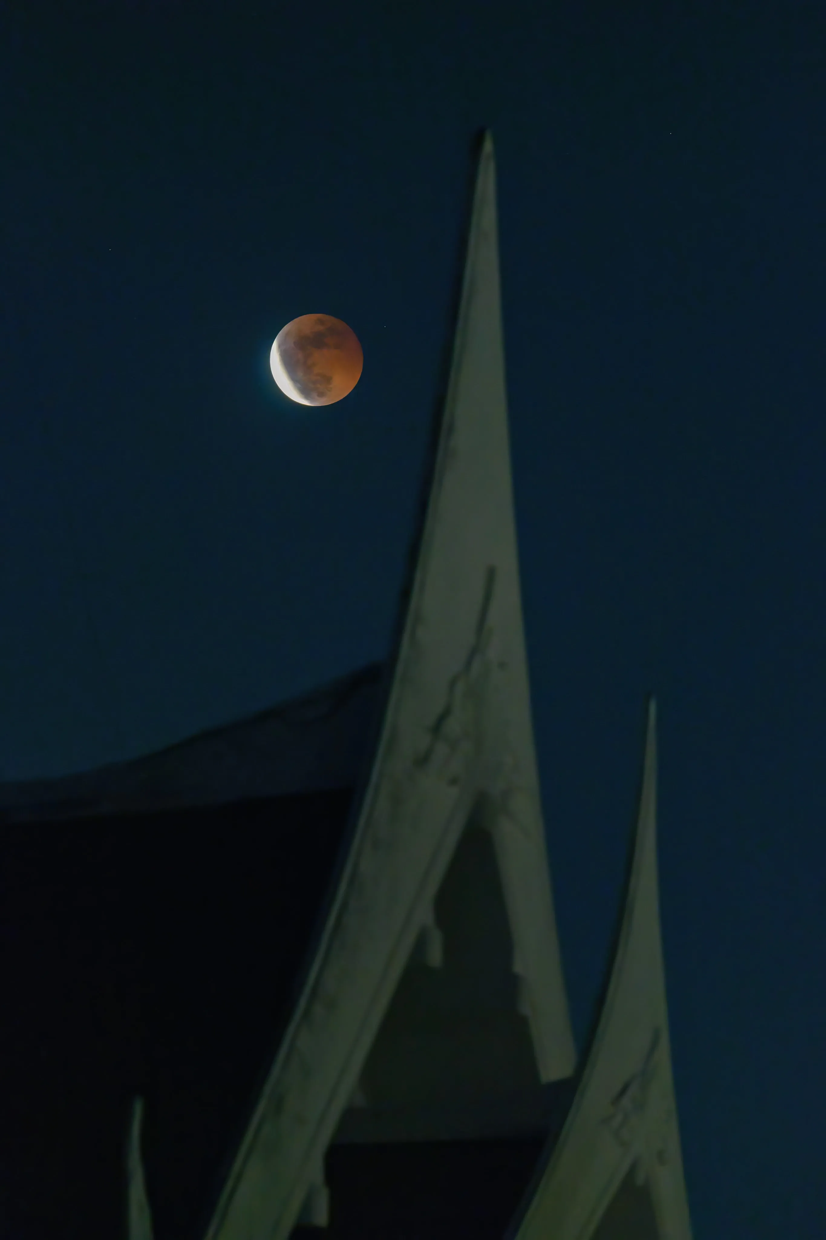 A crescent moon in partial eclipse above the ornate spires of a temple roof at dusk.