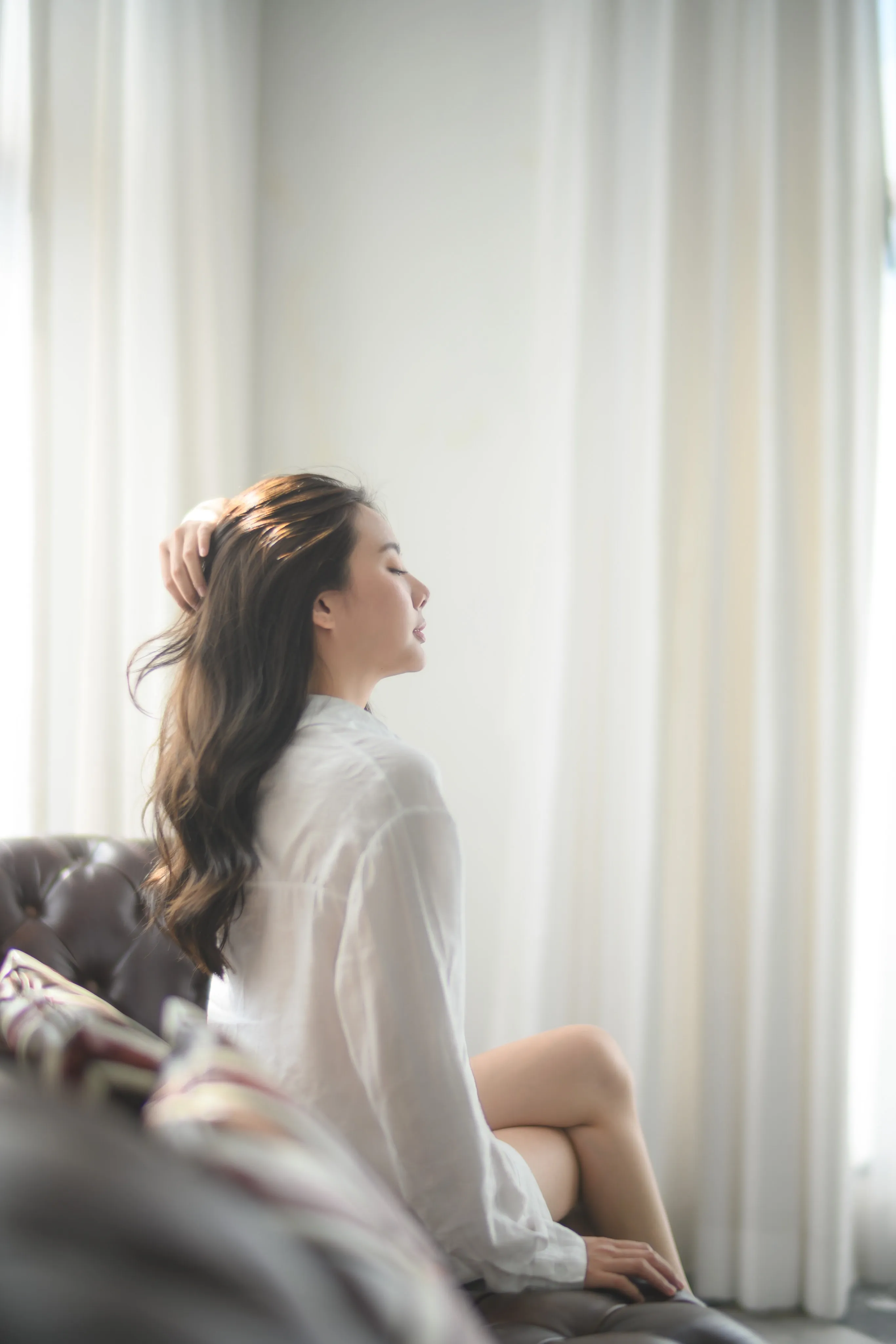 A woman in a white shirt seated by a sunlit window, hair back-lit.