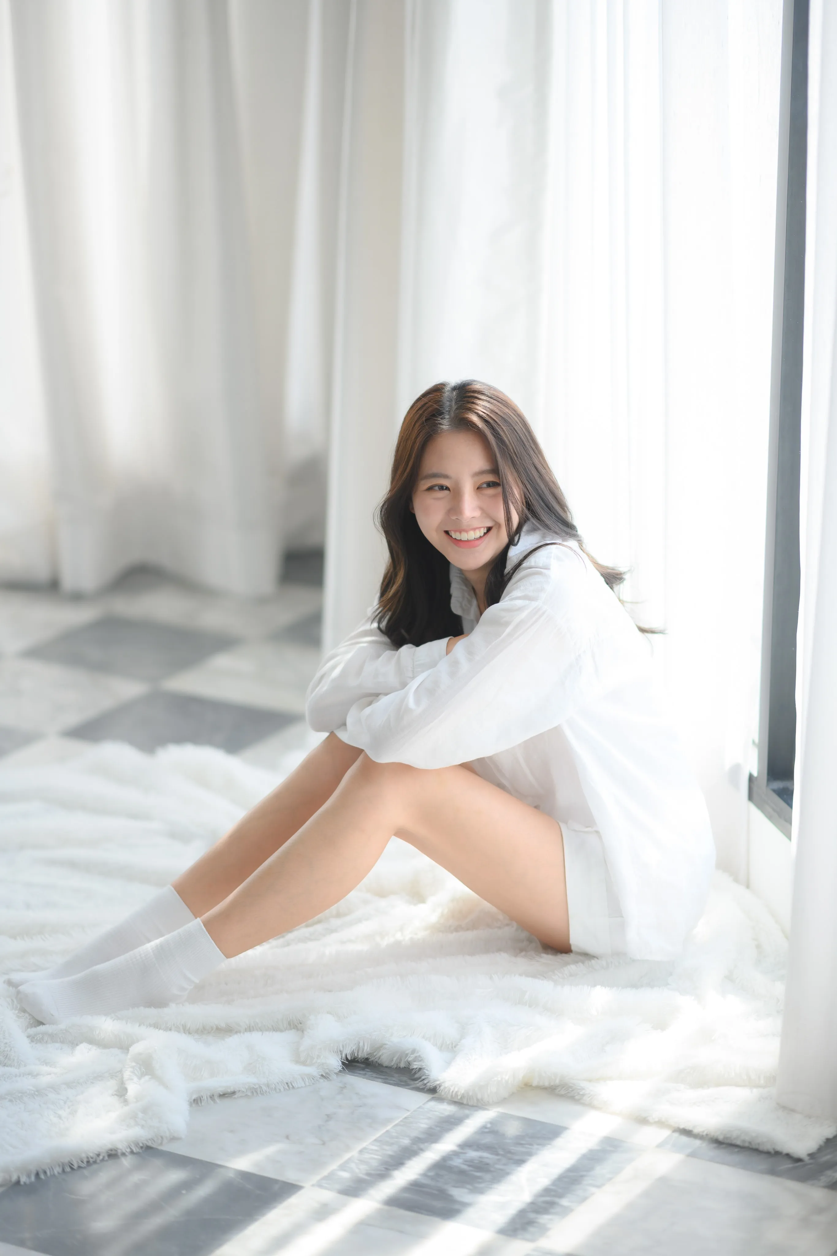 A woman in a white shirt sitting on a fur rug by a bright window.