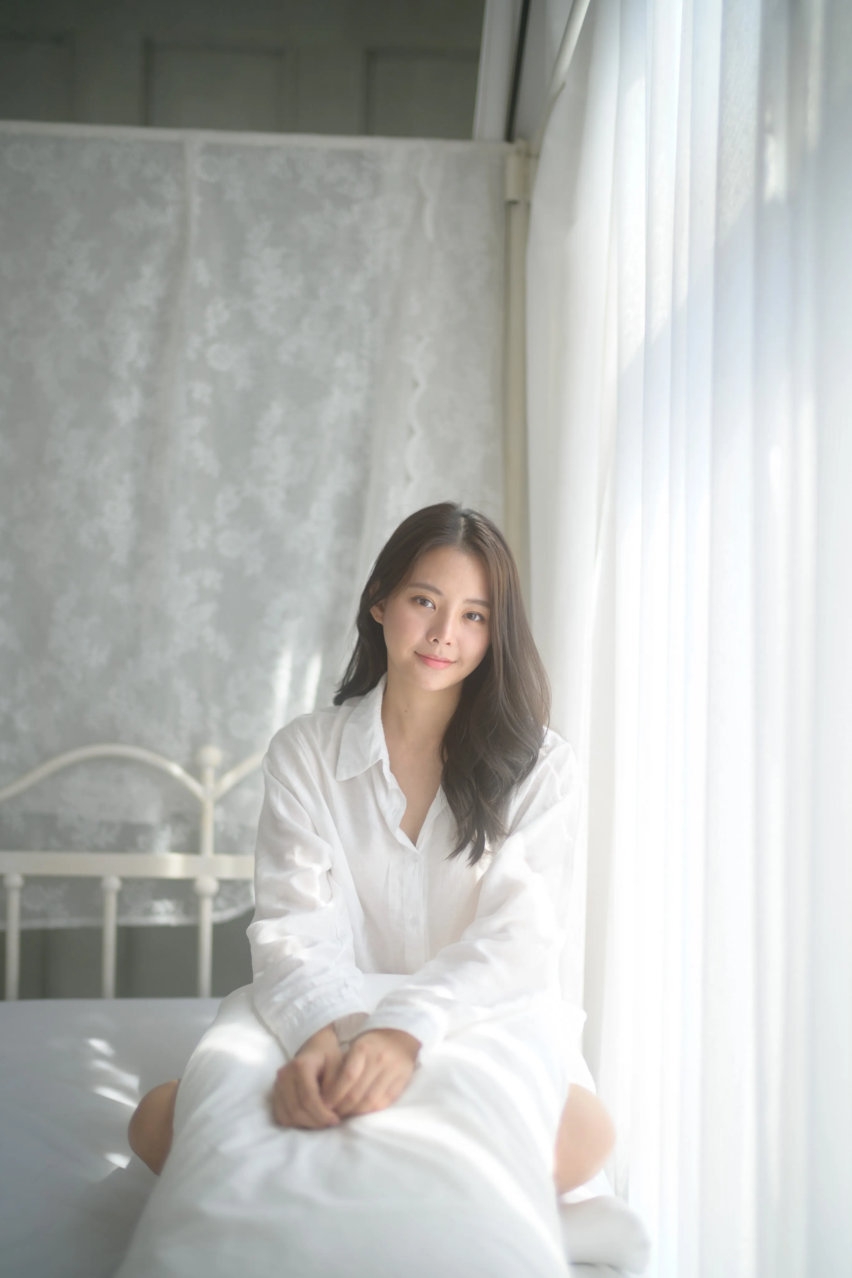 A woman in white loungewear seated on a bed by sheer curtains.