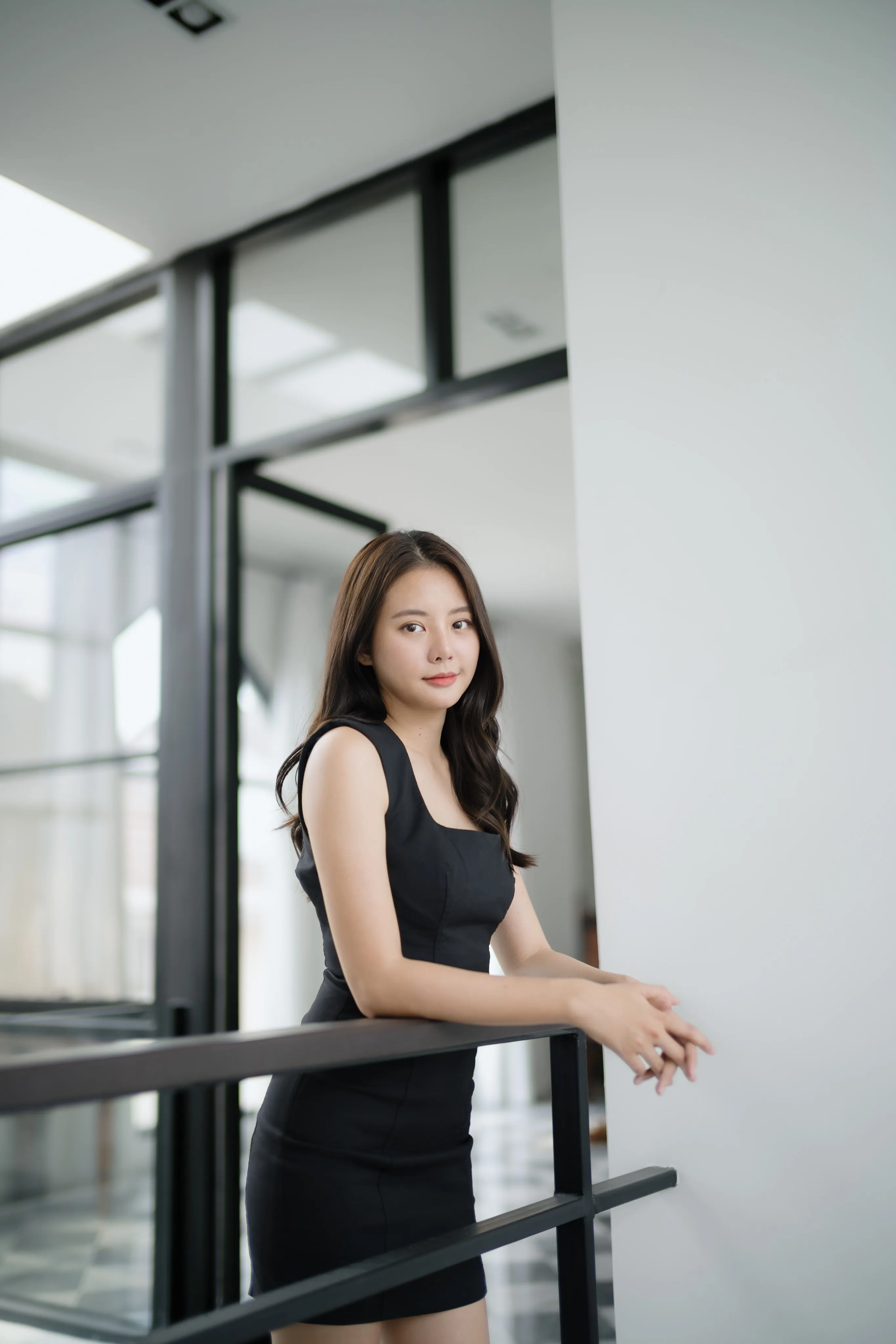 A woman in a black dress leaning against a glass stair railing.
