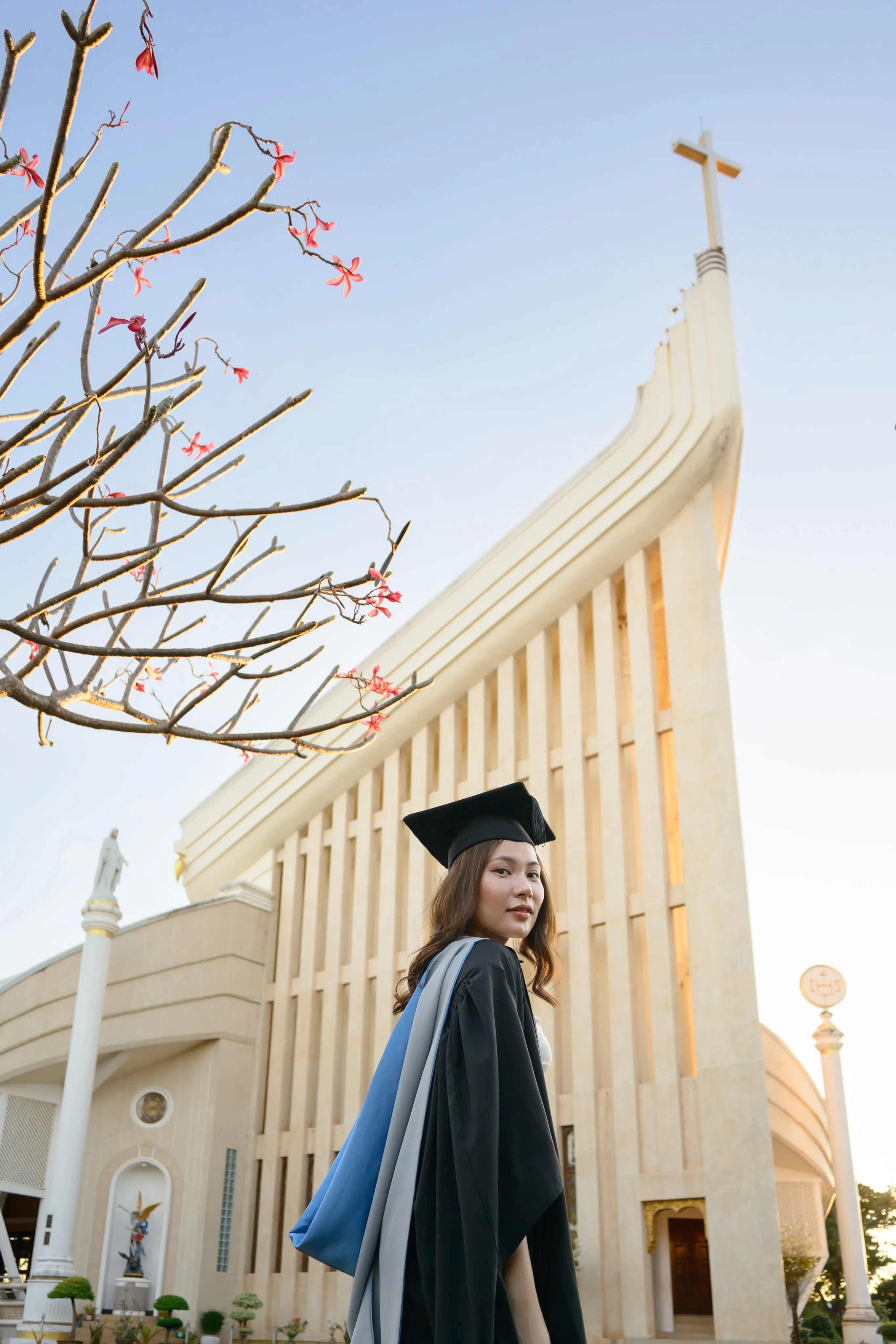 A graduate in cap and gown in front of a white church topped with a cross, framed by blossom branches