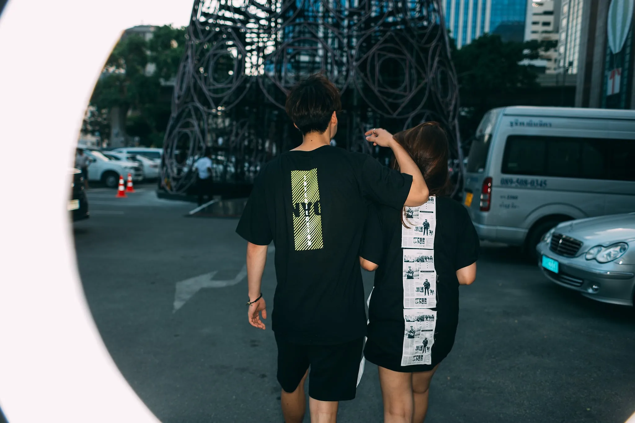 Two people in matching black graphic T-shirts walking away from the camera across a city street, seen through a fisheye vignette