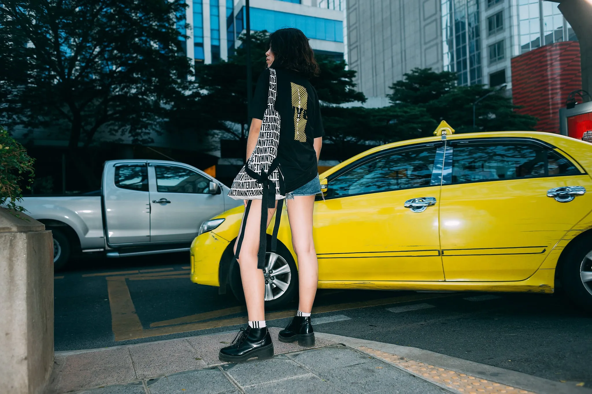 A young woman in a black graphic T-shirt with a patterned tote bag standing beside a yellow taxi on a city street