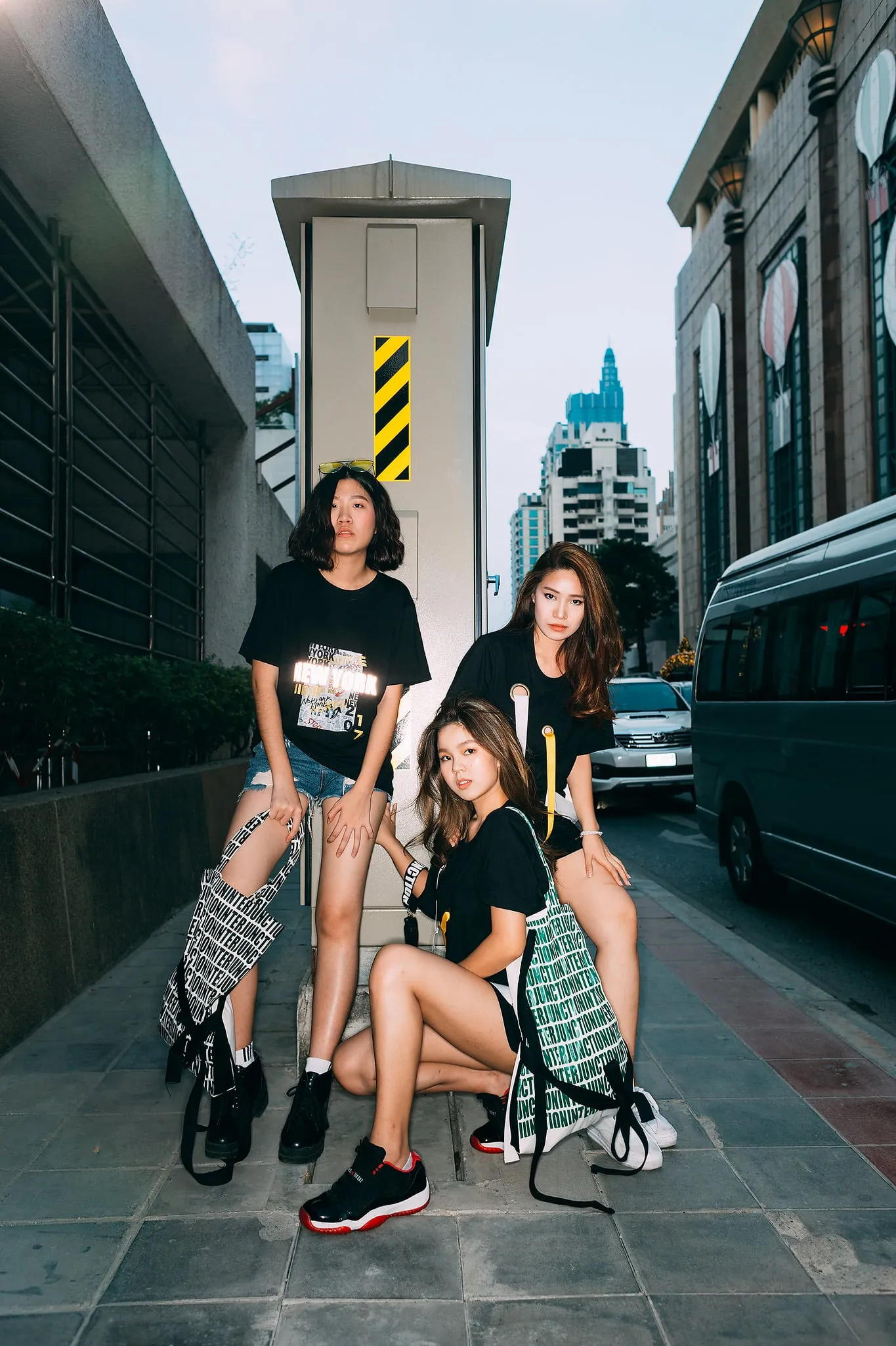Three young women in black graphic T-shirts with patterned tote bags posing together on a city sidewalk