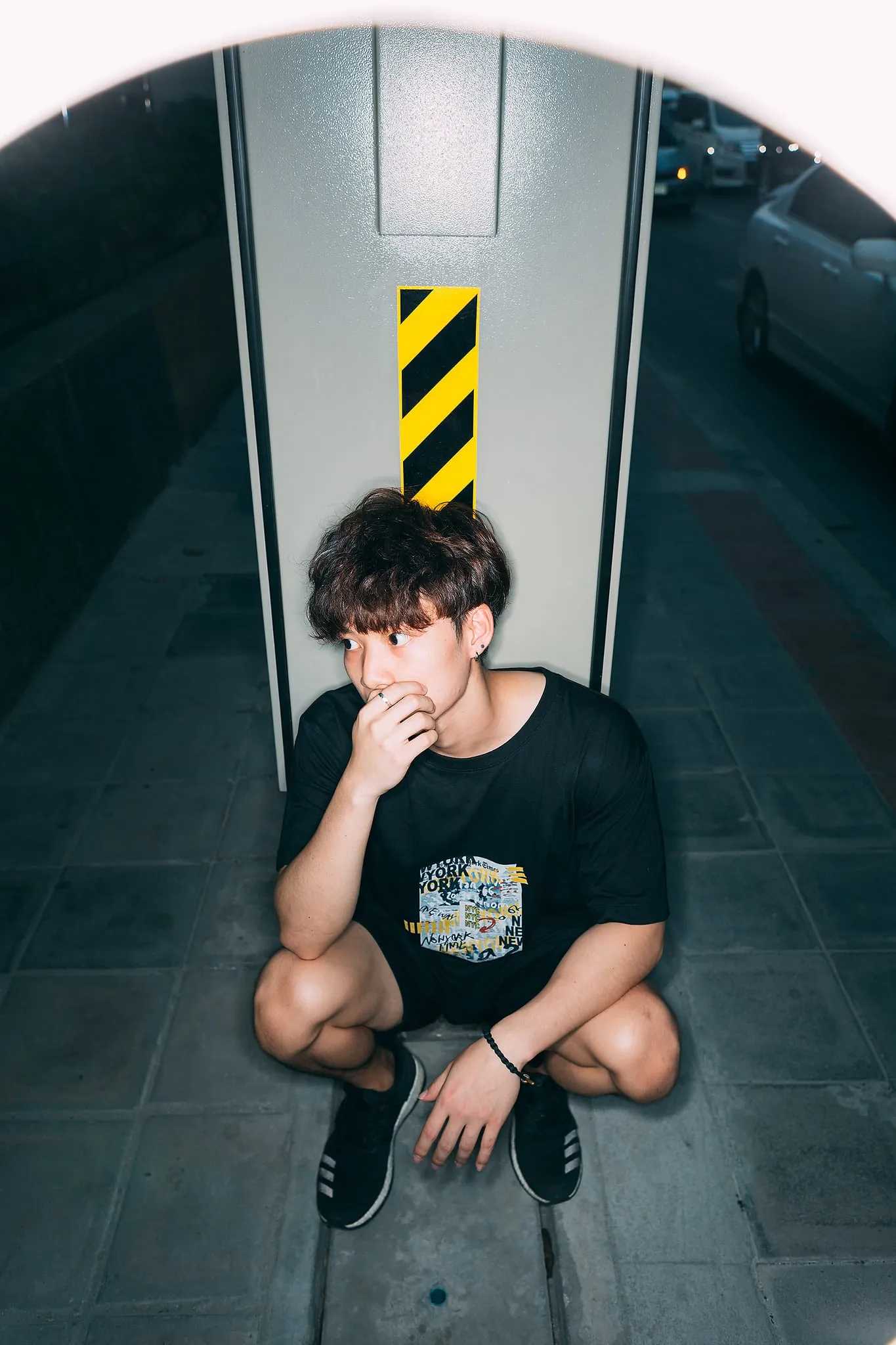 A young man in a black graphic T-shirt crouching against a pillar marked with yellow-and-black hazard stripes