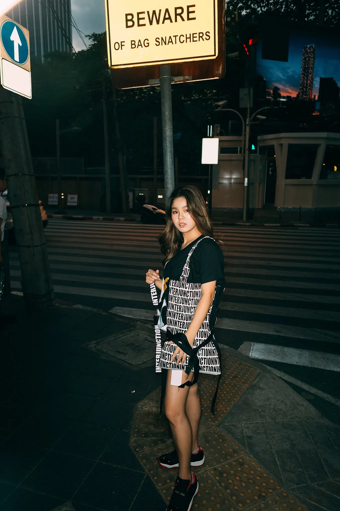 A young woman in a black T-shirt with a patterned tote bag at a street crossing under a "Beware of bag snatchers" sign
