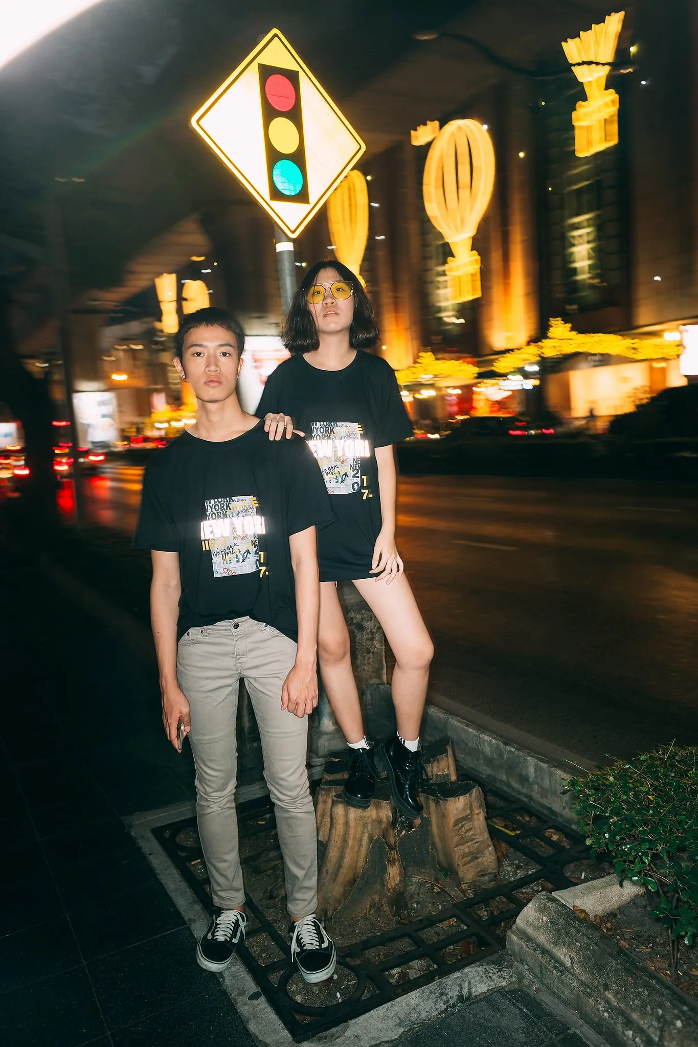 Two young people in black graphic T-shirts standing on a city sidewalk at night beneath glowing hot-air-balloon lights