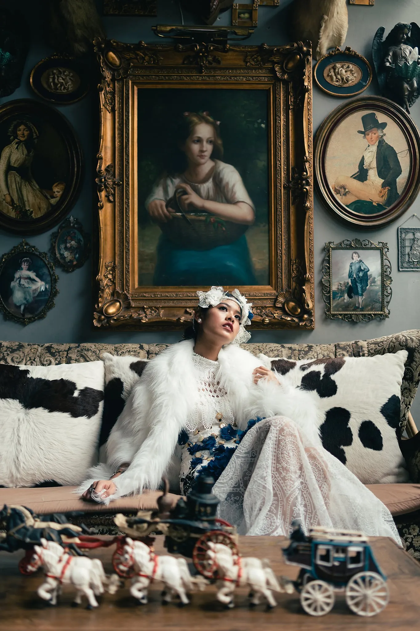 A woman in a white fur coat and lace gown reclining on a cowhide sofa beneath gilded antique portraits