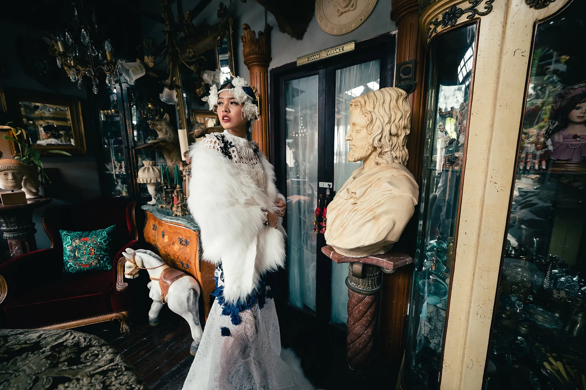 A woman in a white fur stole and lace gown standing among antiques beside a marble bust