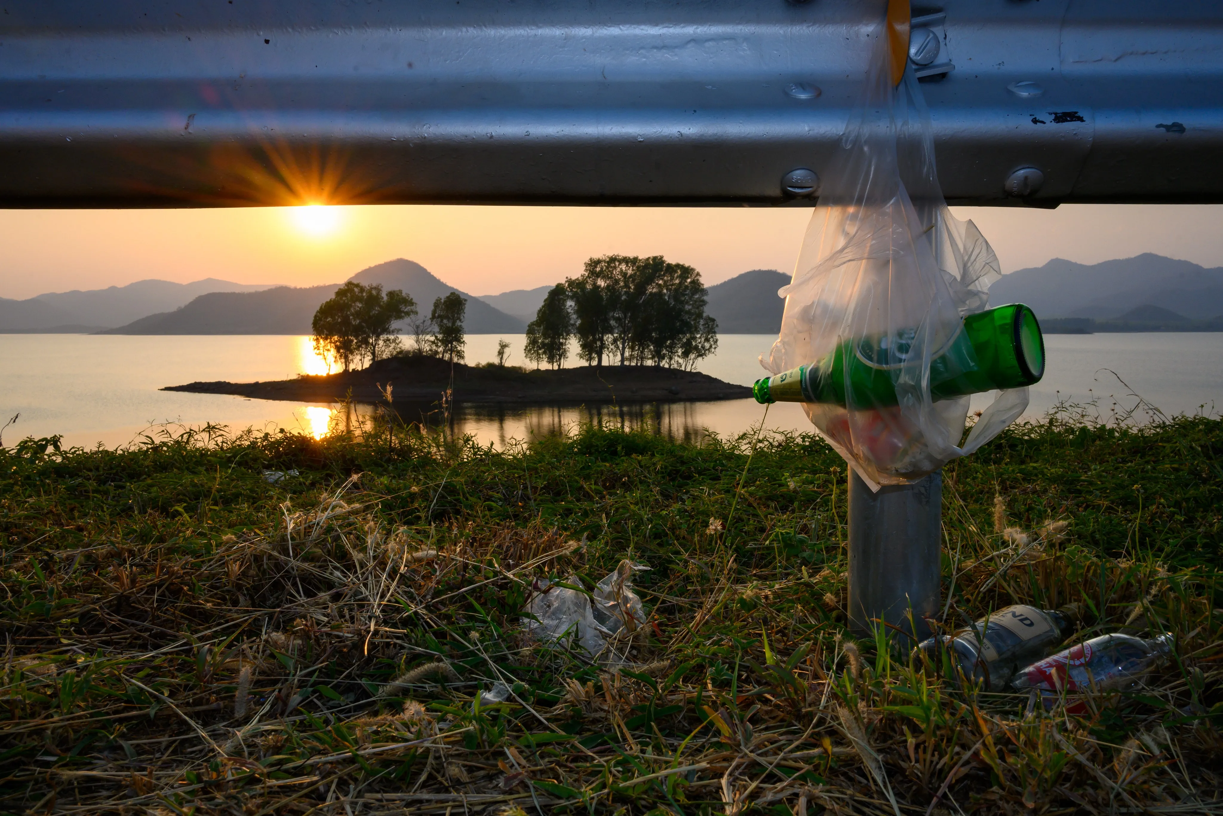 A discarded plastic bag and bottle caught on a guardrail, silhouetted against an island sunset.