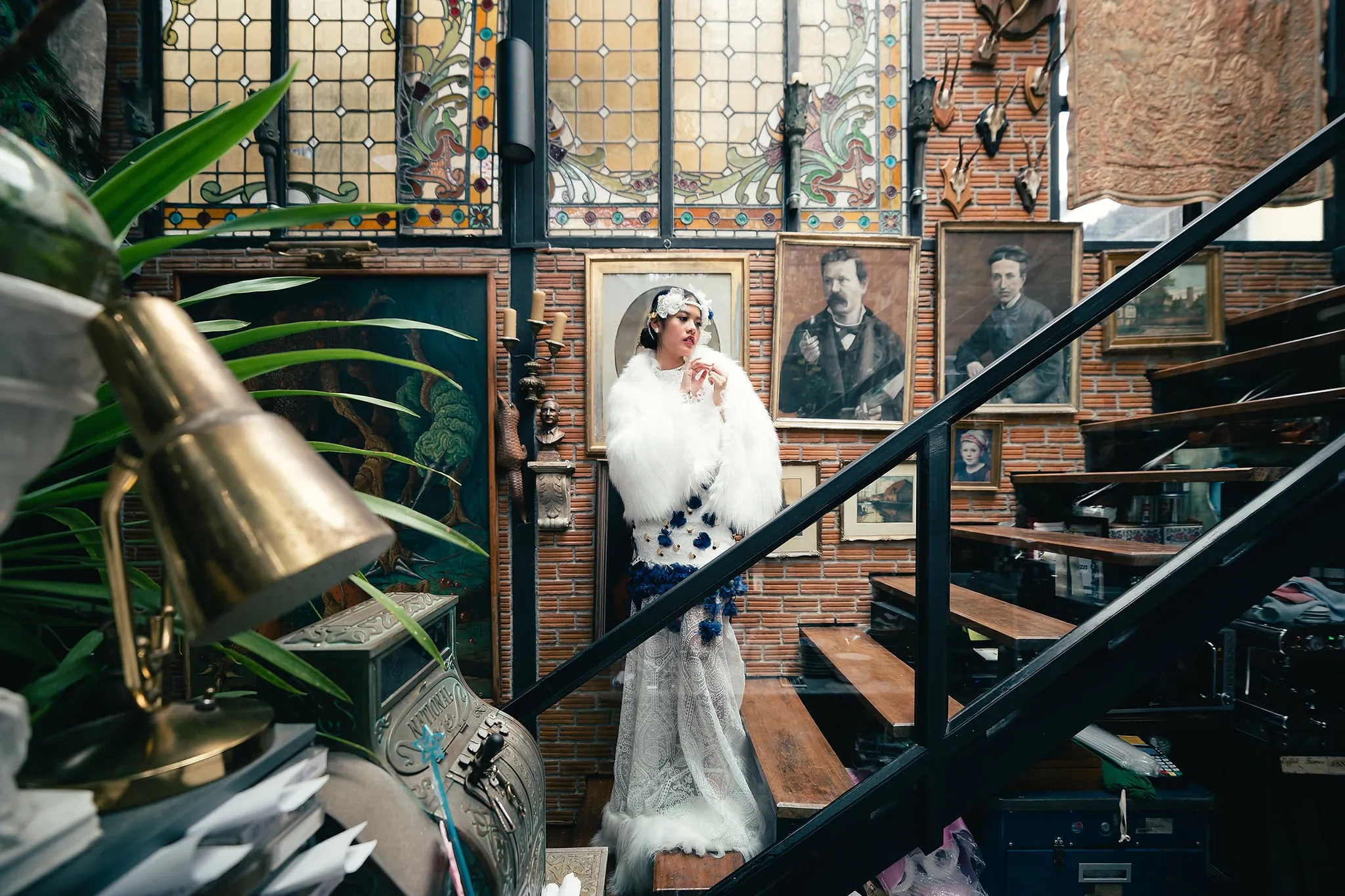 A woman in a white fur coat and lace gown on a staircase in a brick room hung with vintage portraits
