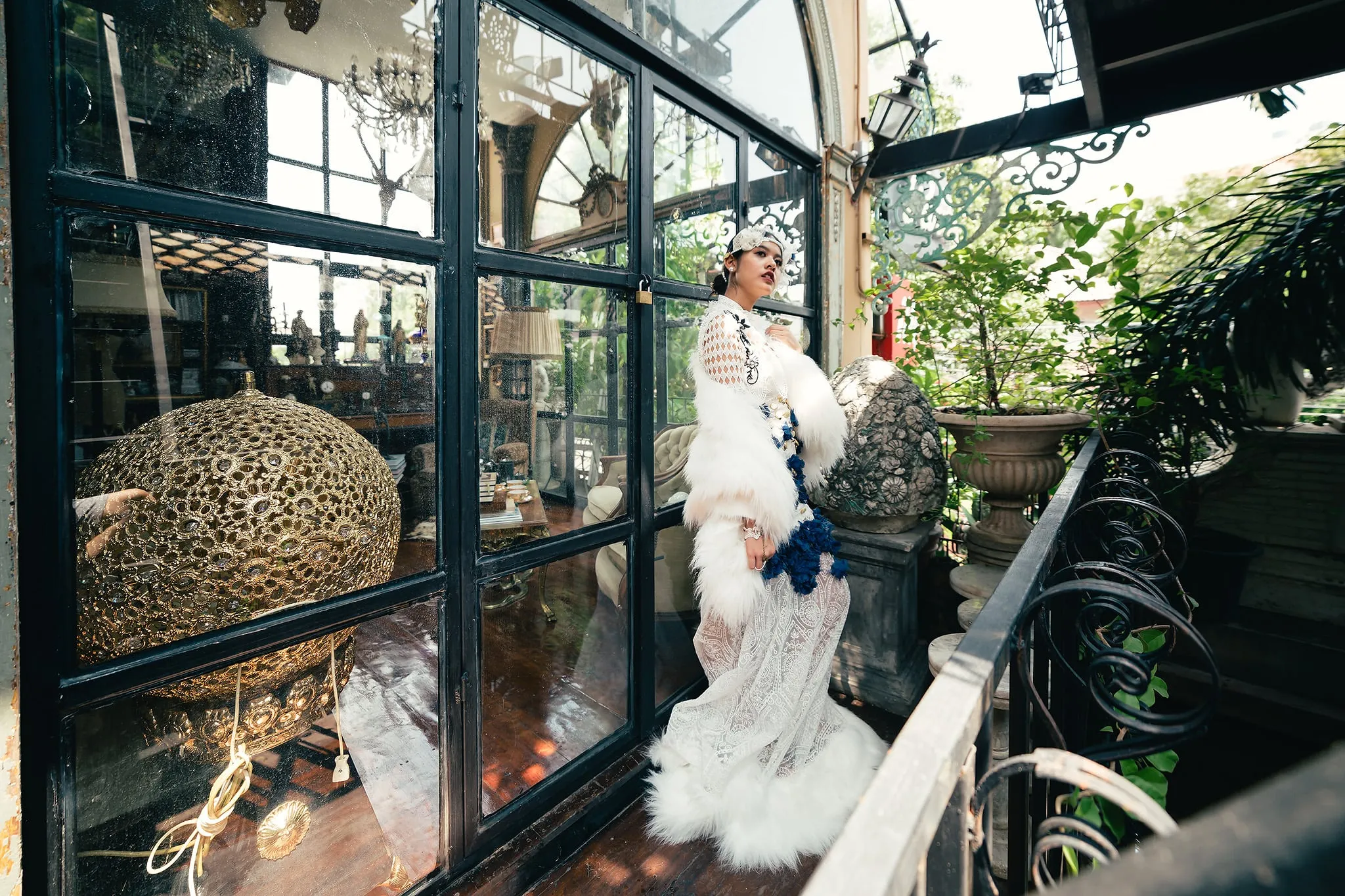 A woman in a white fur coat and lace gown posing in a glass conservatory filled with antiques and plants