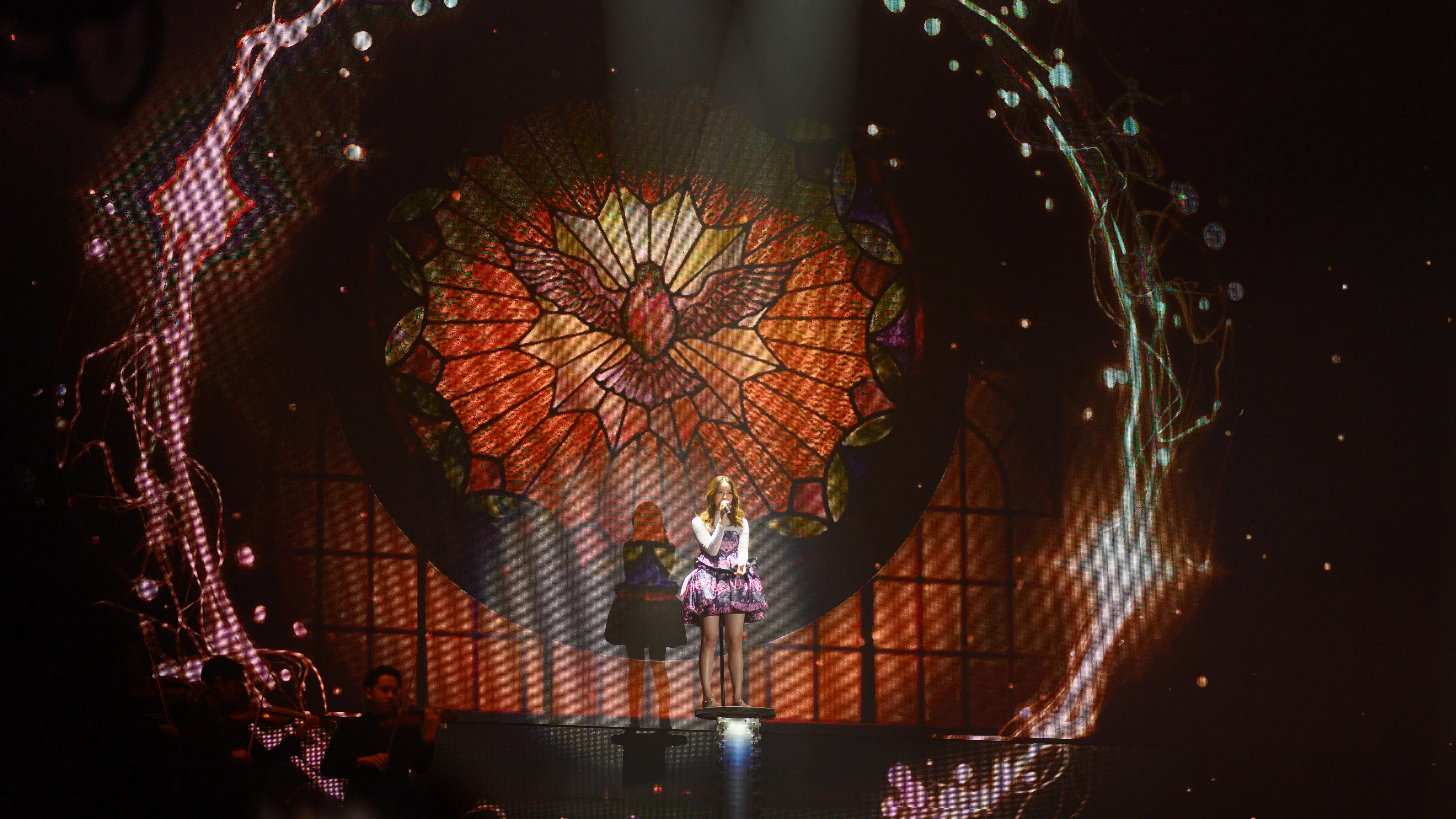 A singer in a purple dress performing on stage in front of a glowing stained-glass dove backdrop