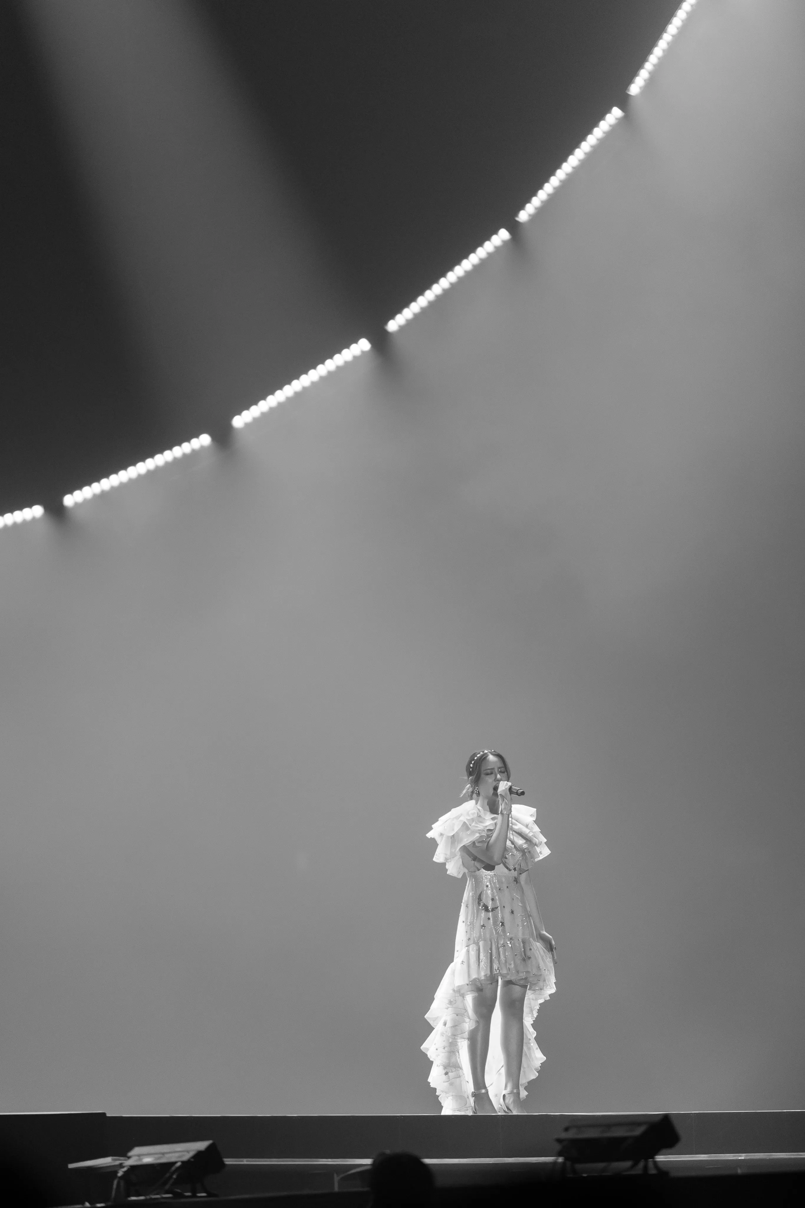 A black-and-white photograph of a singer in a ruffled white dress on a stage beneath a curved lighting rig