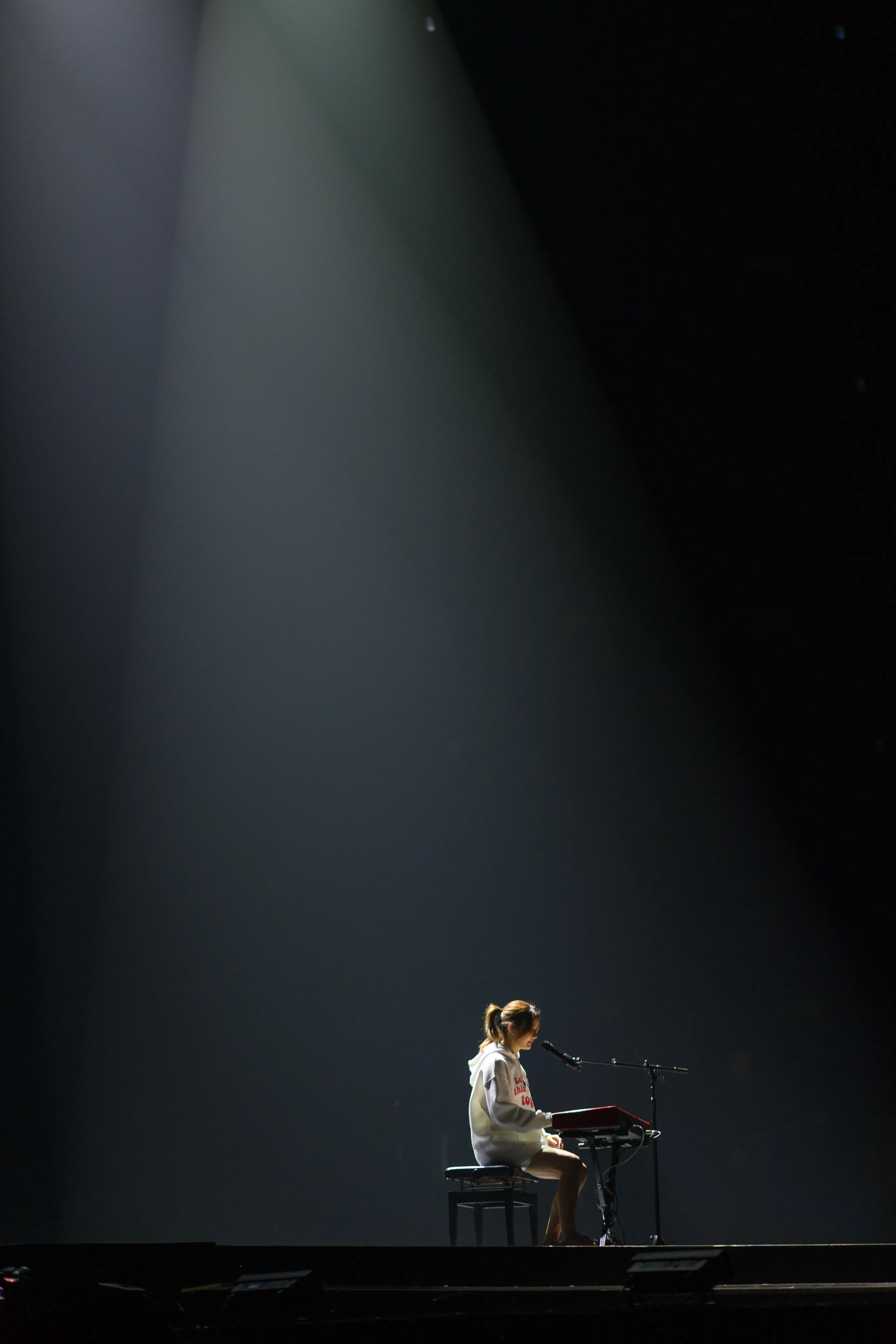 A singer seated at a keyboard alone on a dark stage beneath a single beam of light