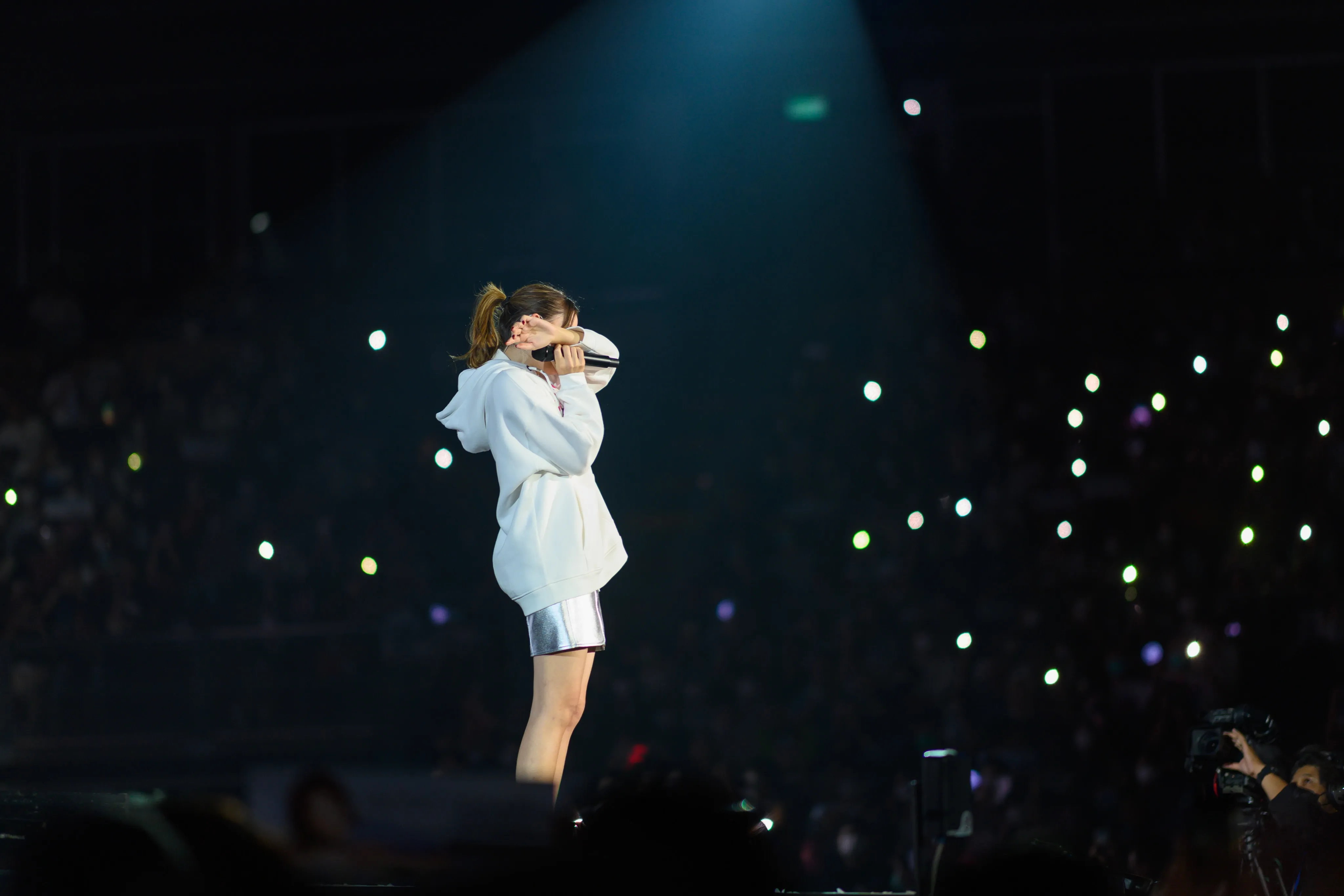 A singer in a white hoodie on stage, hands to her face, before an arena of glowing light sticks