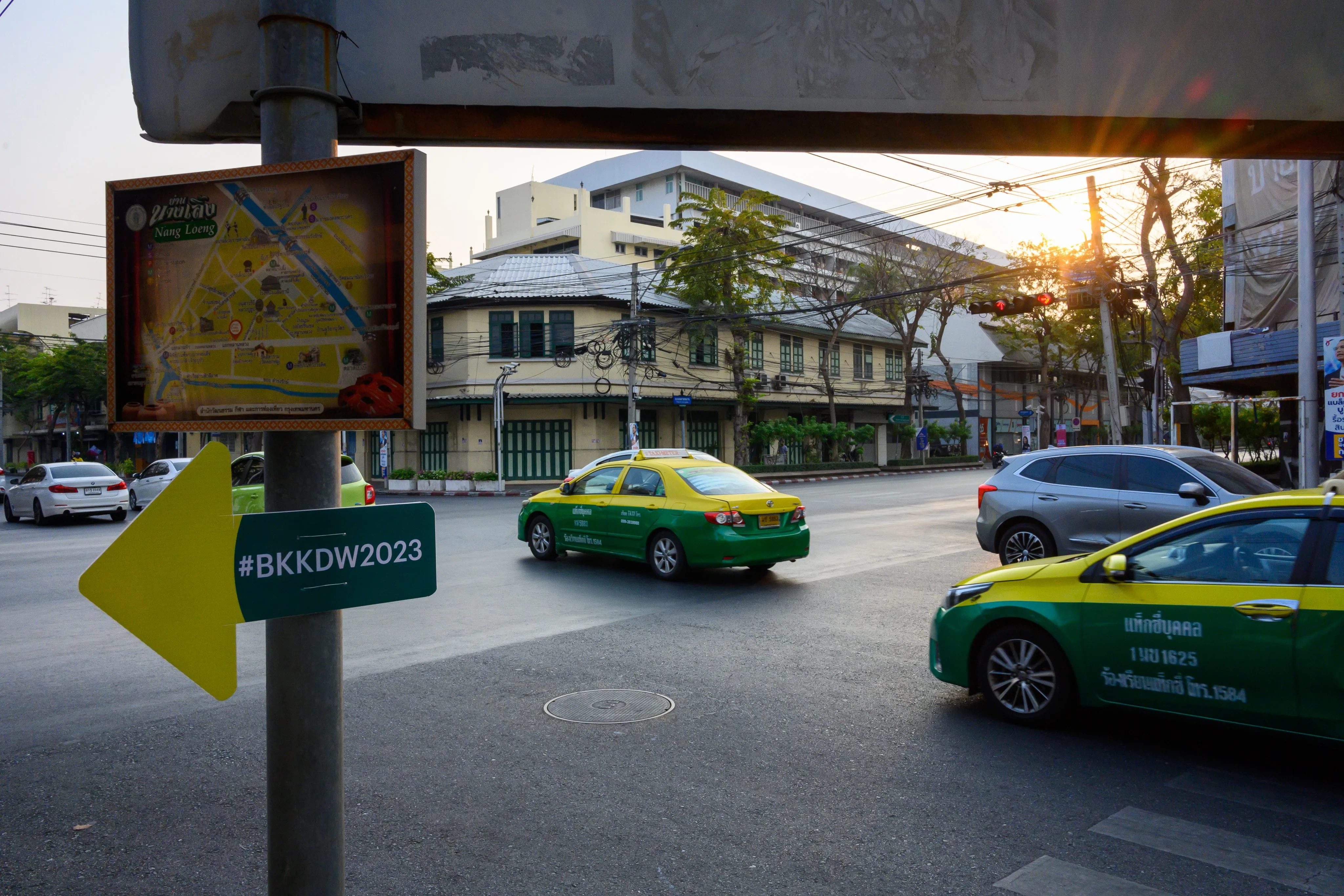 A Bangkok Design Week wayfinding sign on a street corner, with taxis passing by.