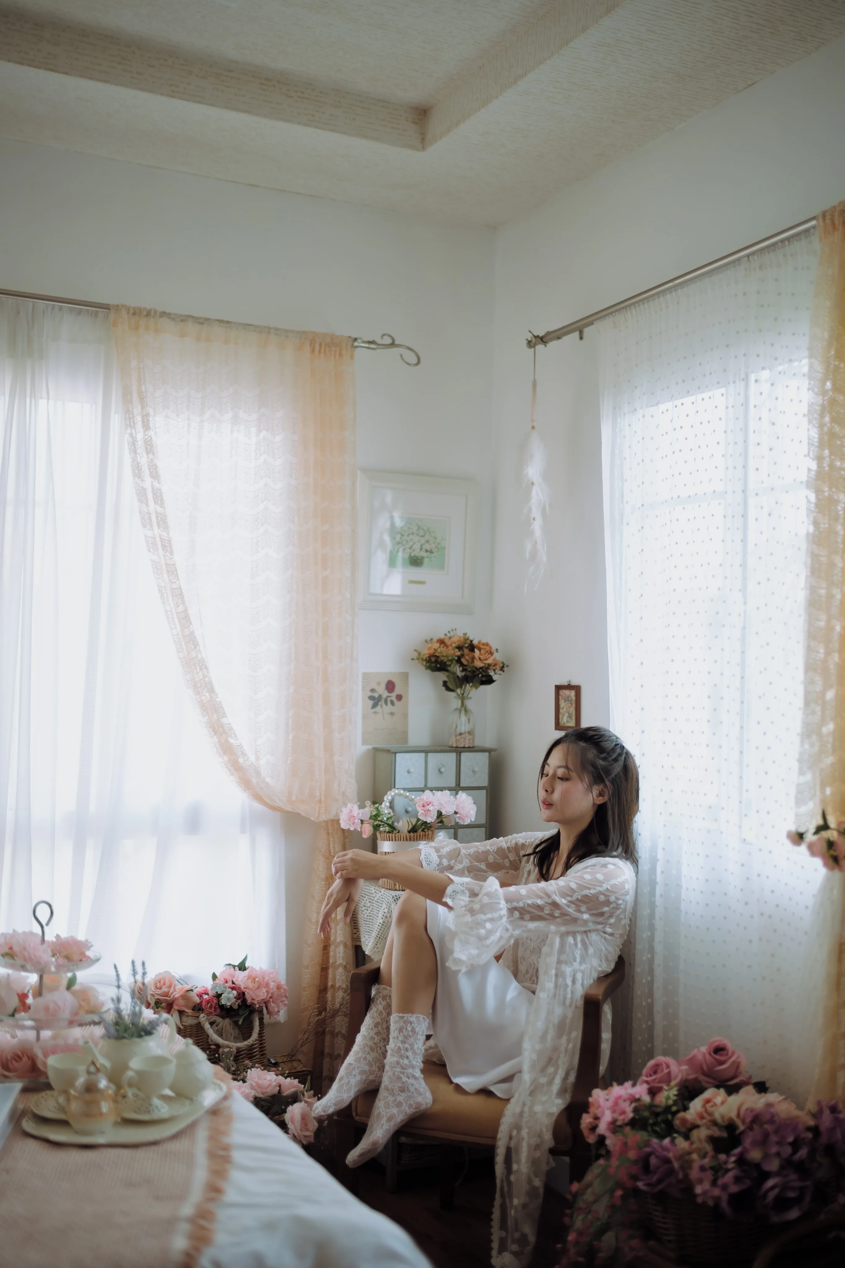 A woman in white lace lounging on a window seat in a sunlit, flower-filled room.