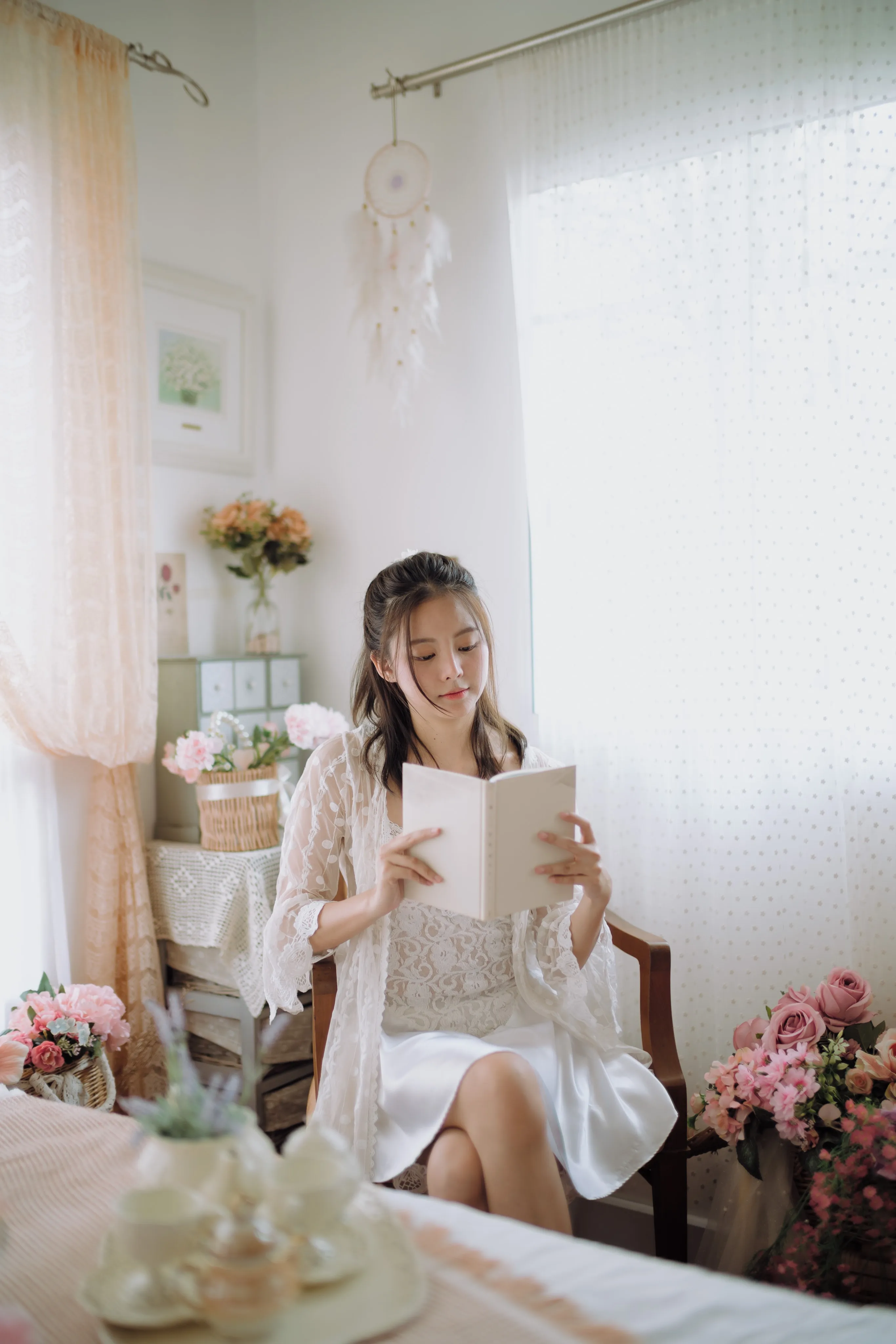 A woman in white reading a book in a sunlit room surrounded by pink flowers.