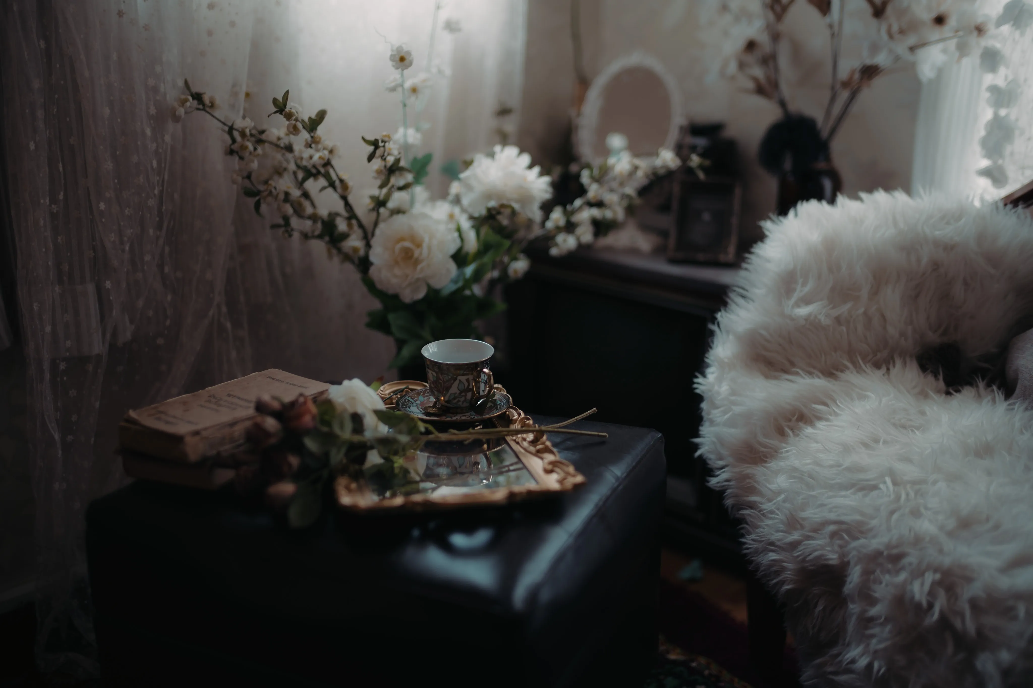 A dark still life of a tea set, books, and flowers on a shadowed side table.