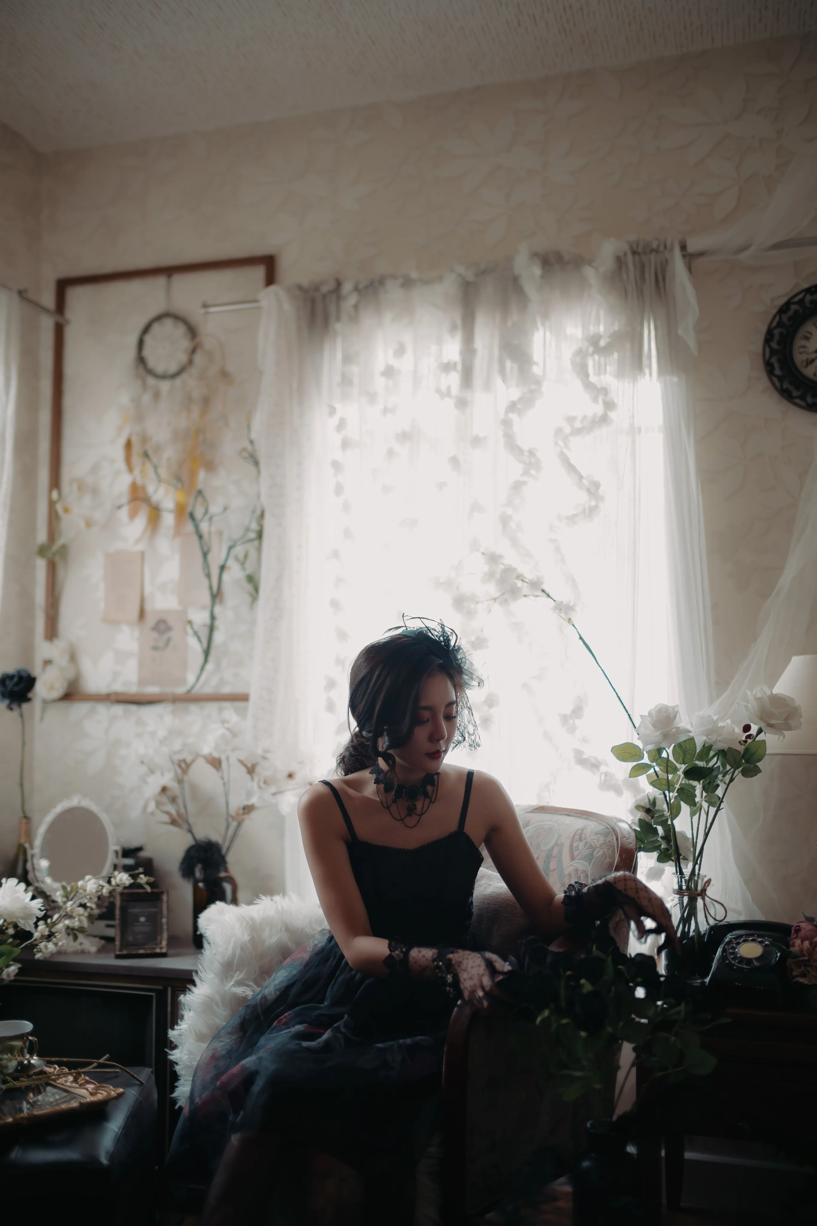 A woman in a black dress seated among dark furnishings, dimly lit.