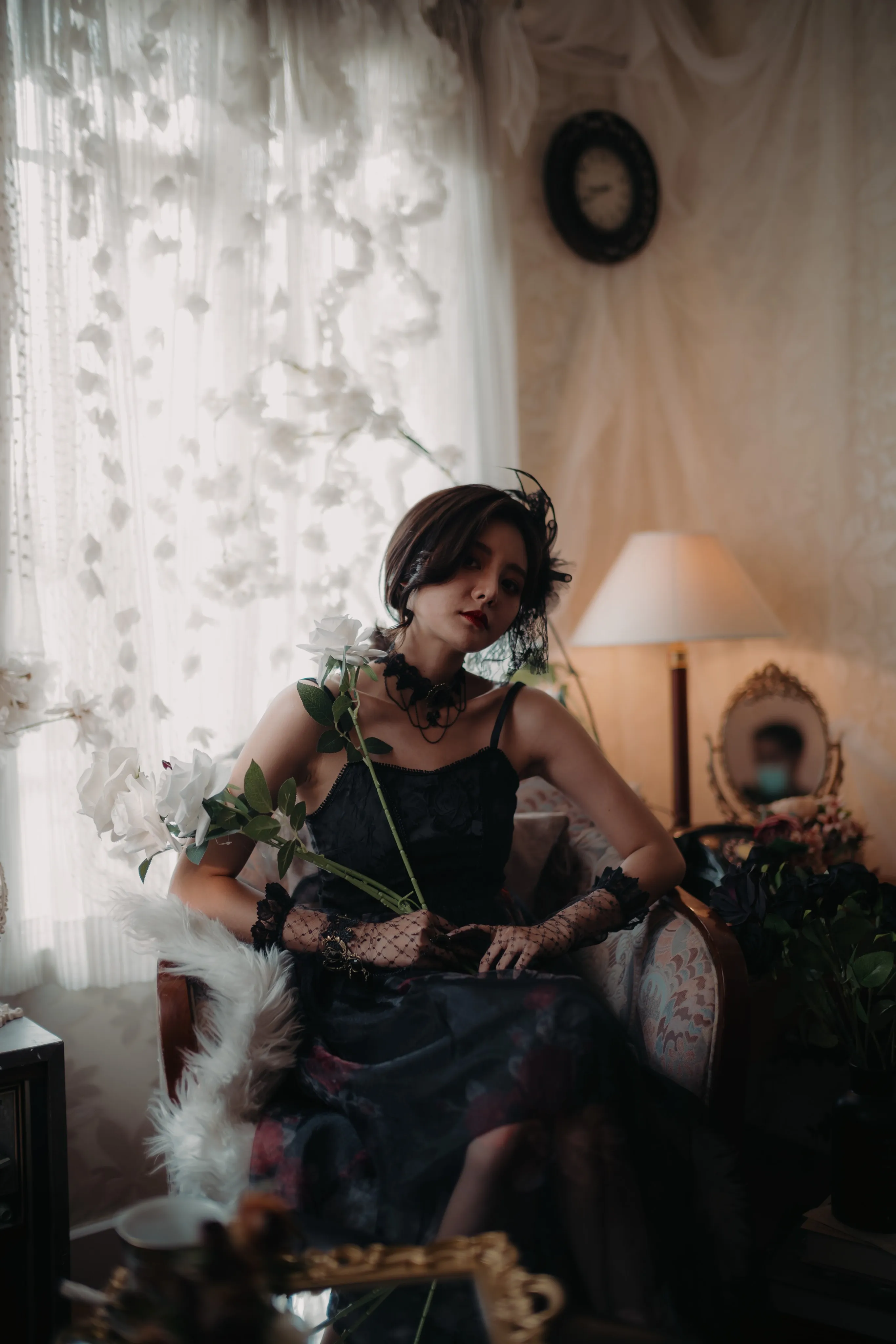 A woman in a black lace dress seated by a sheer curtain in low light.