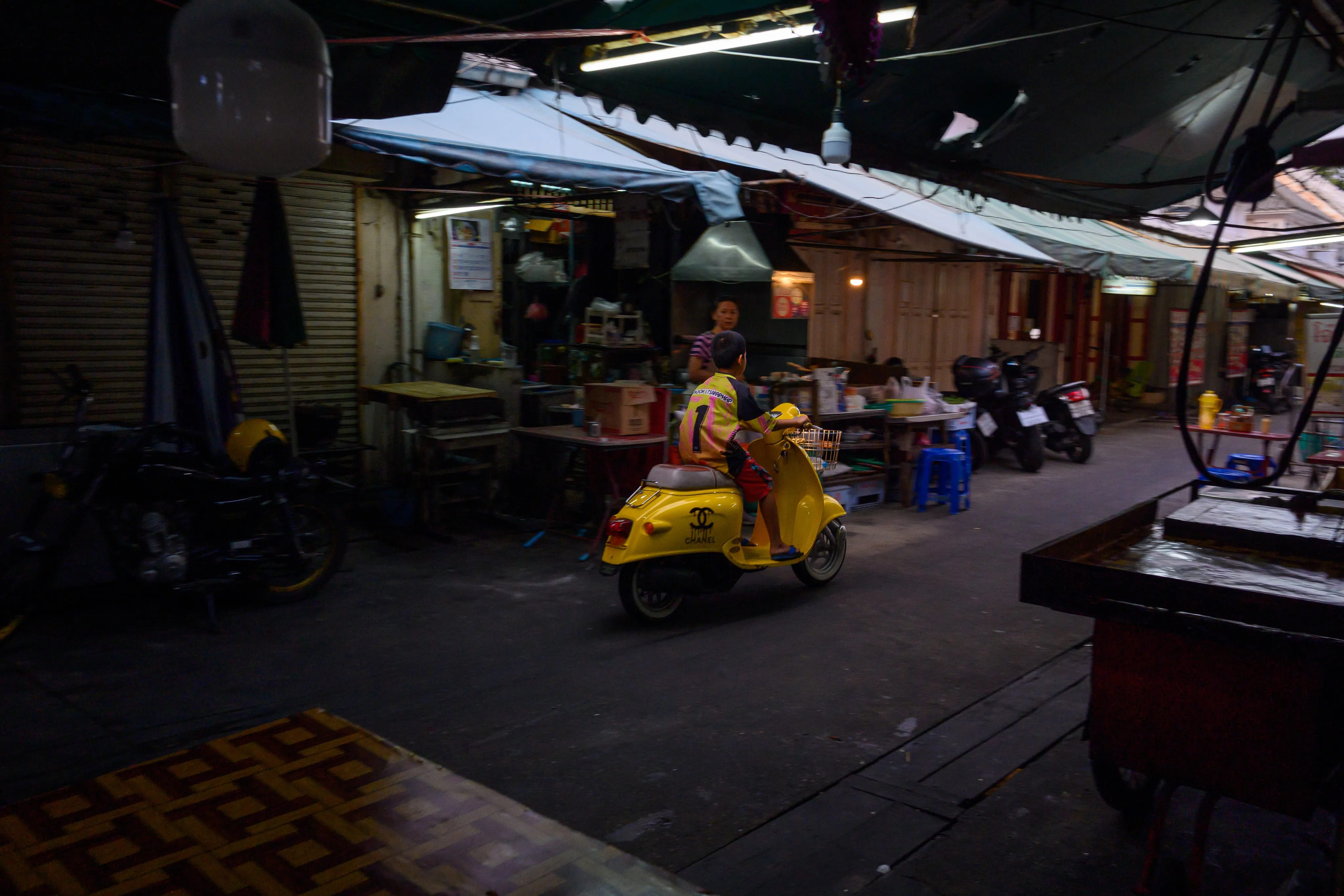 A woman tending a night market food stall beside a parked scooter.