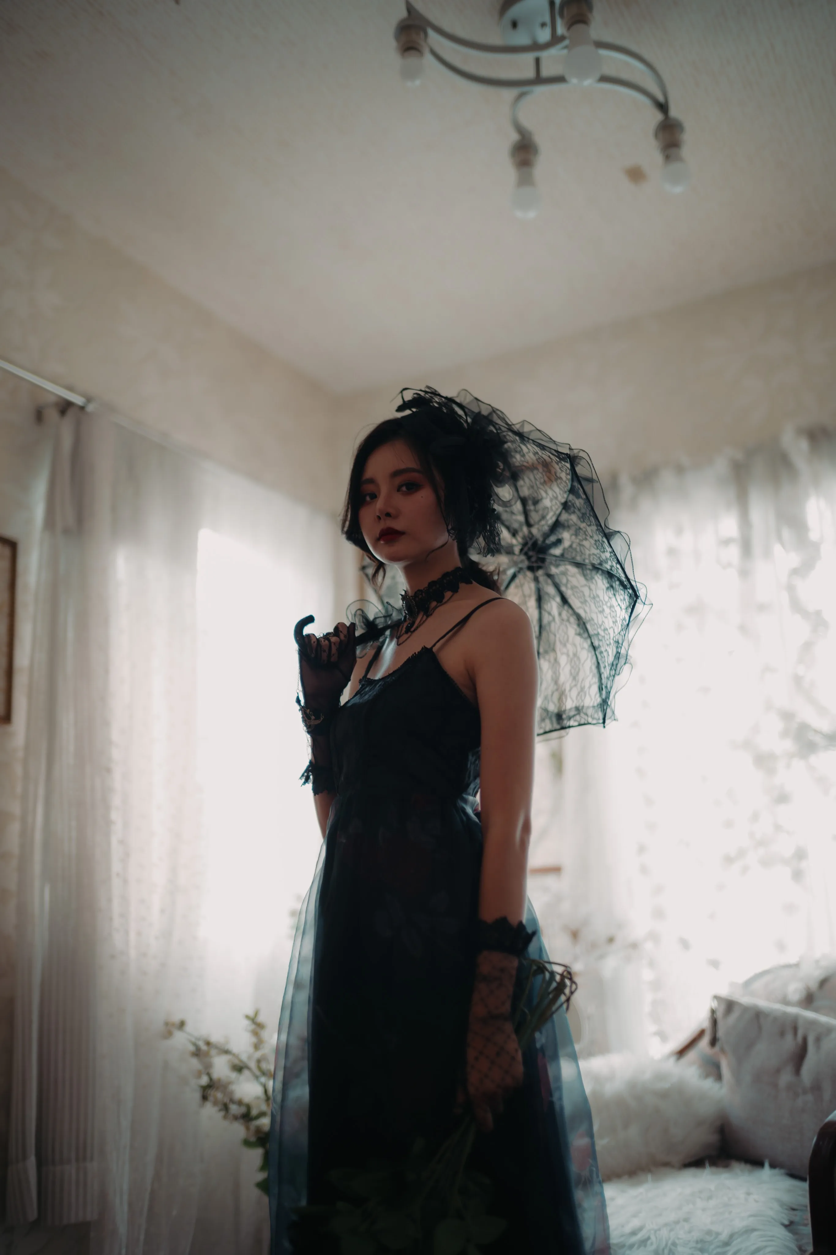 A woman in a black dress holding a lace umbrella, backlit by a window.