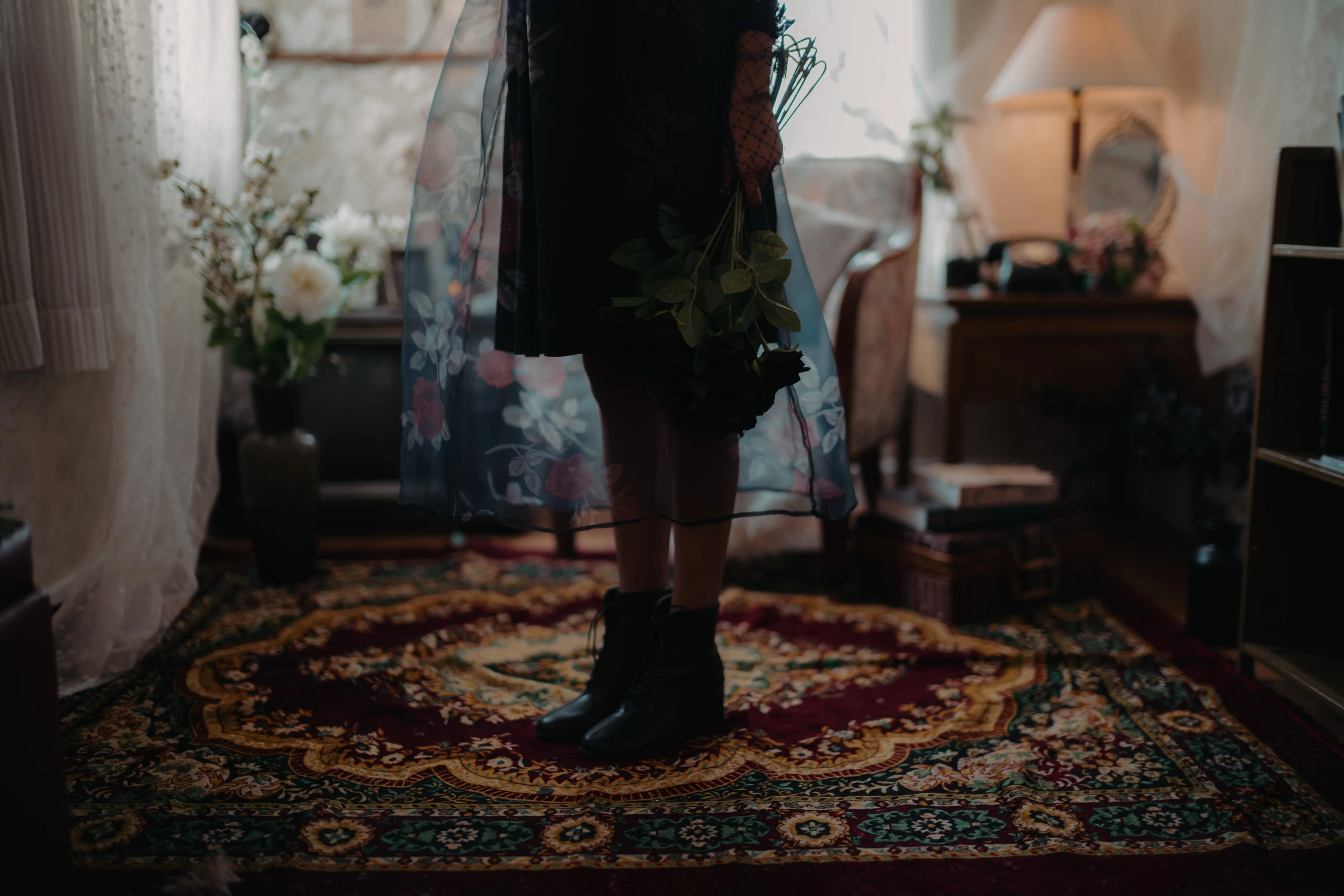A woman in a sheer black dress standing on a patterned rug in a dim room.