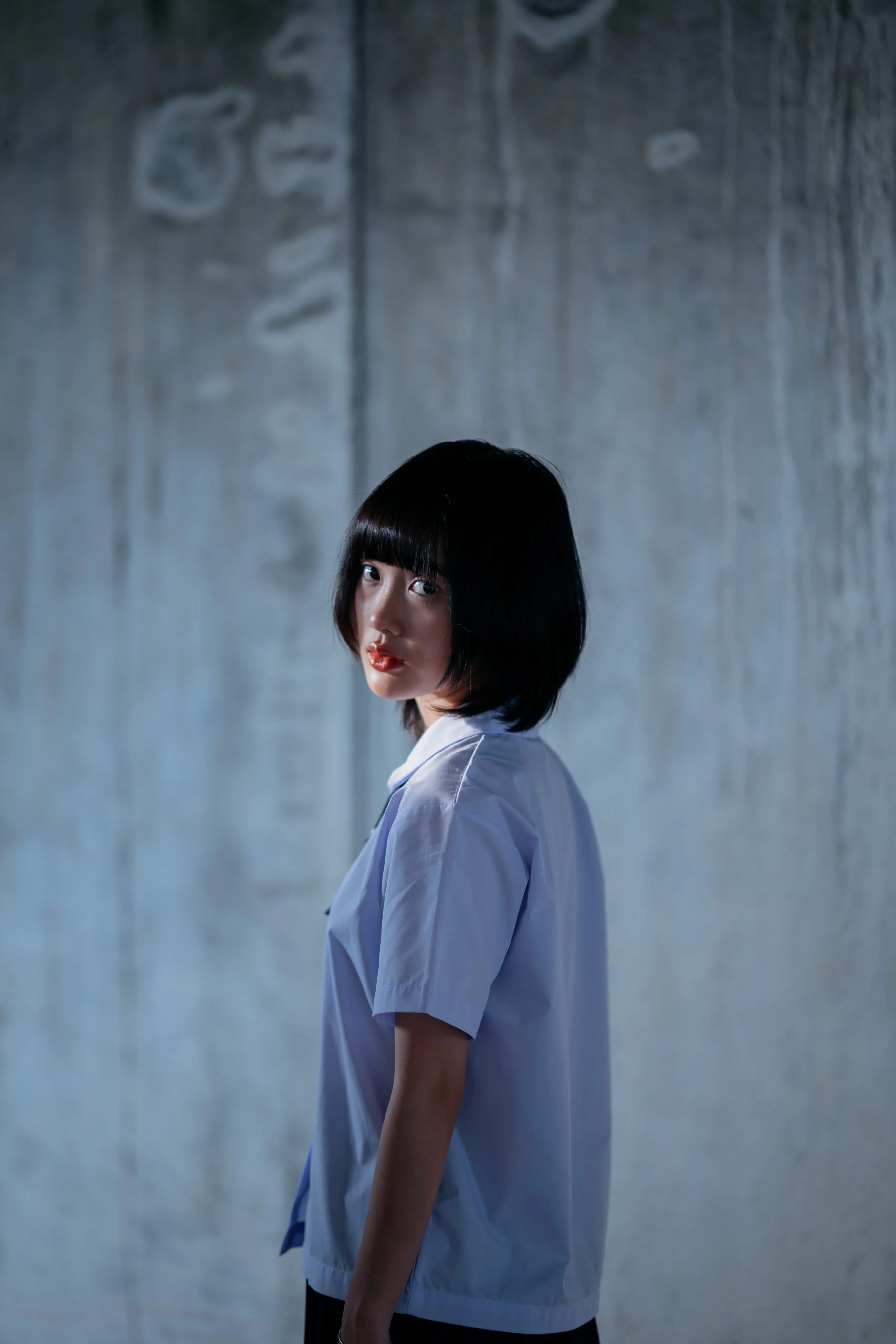 A girl with a bob haircut in a school uniform looking back over her shoulder in a concrete stairwell.