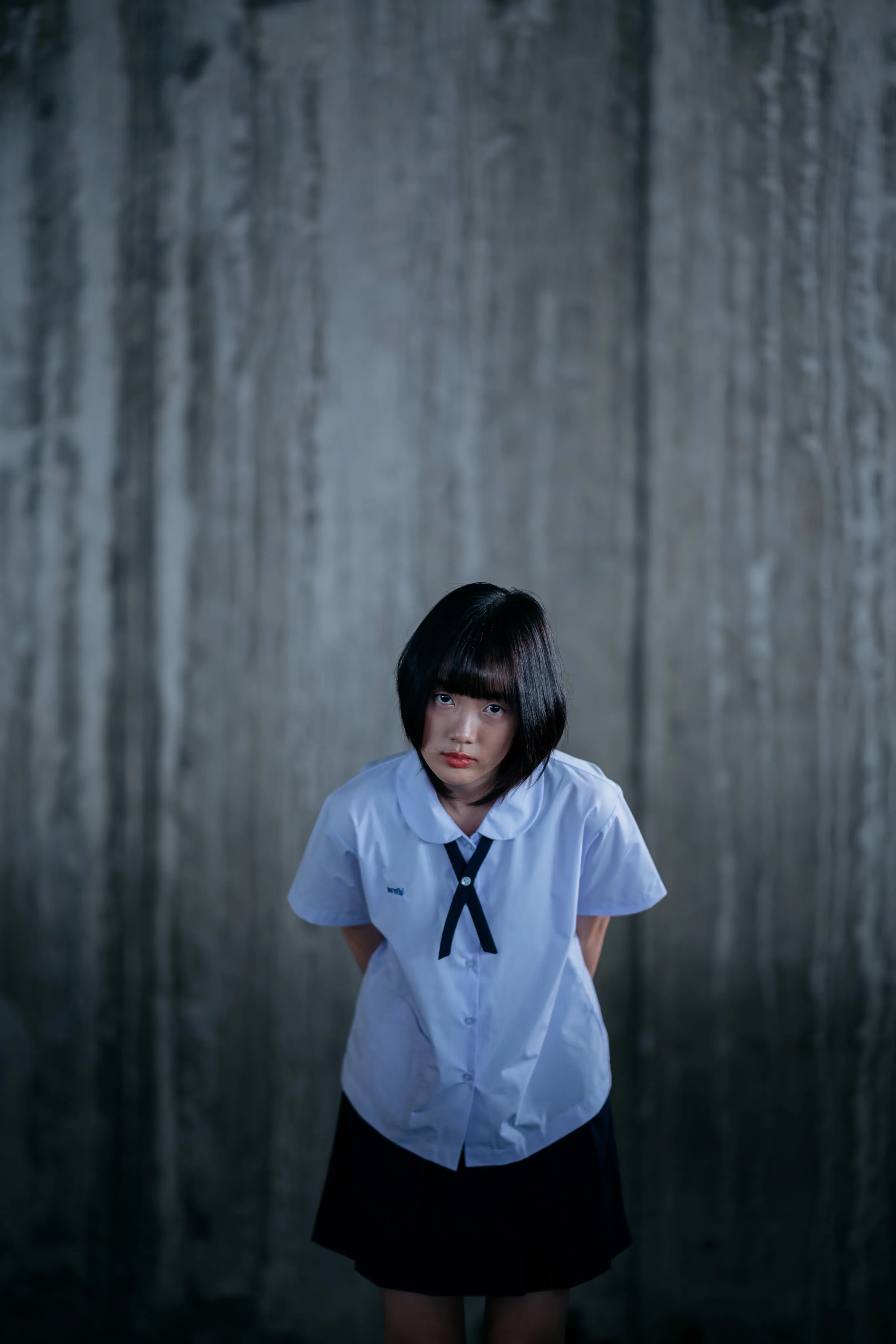 A girl in a school uniform standing with hands behind her back in a dim concrete stairwell.
