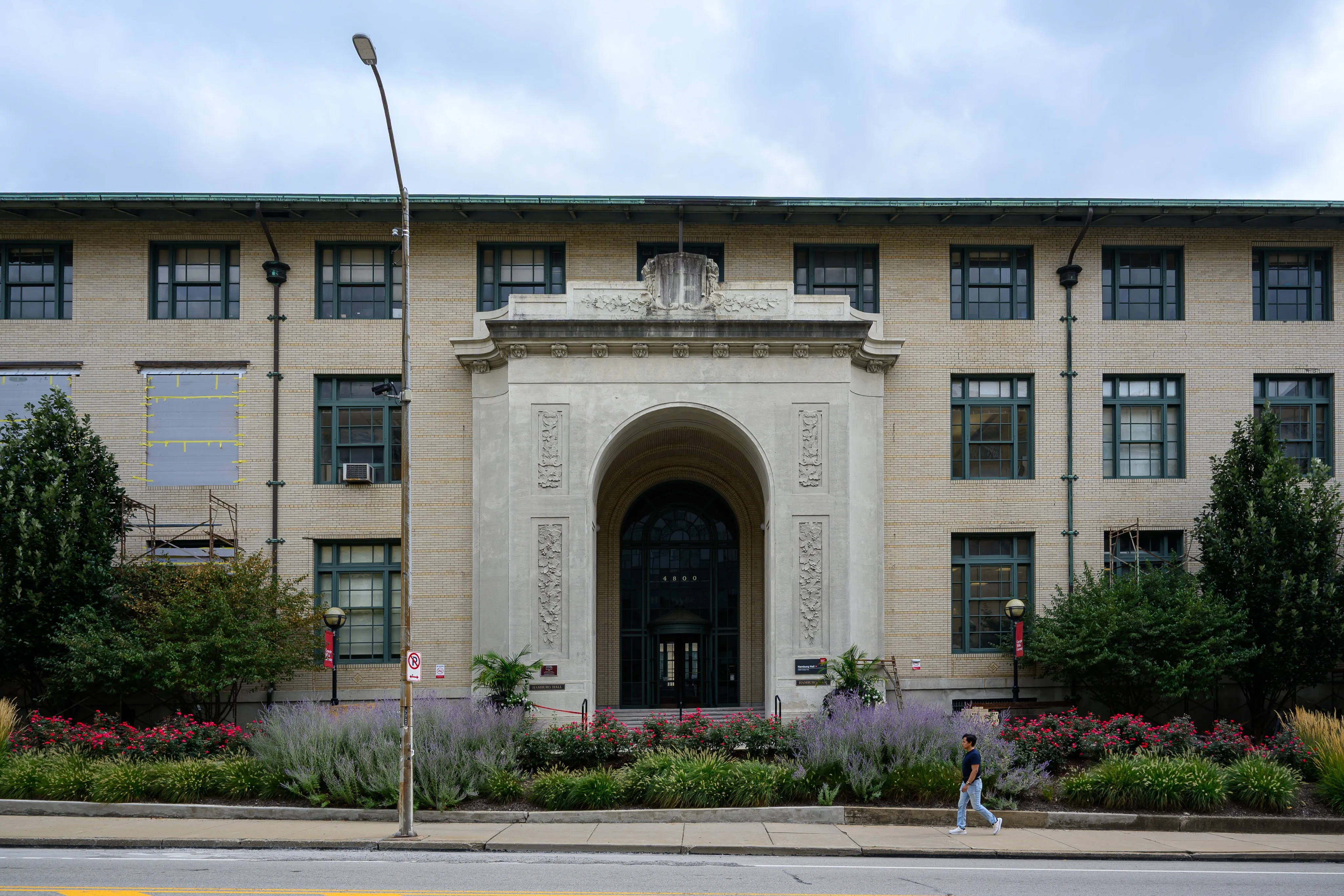A tan brick academic building with a carved stone arched entrance, fronted by beds of lavender and red flowers, with a pedestrian walking past on the sidewalk under an overcast sky.
