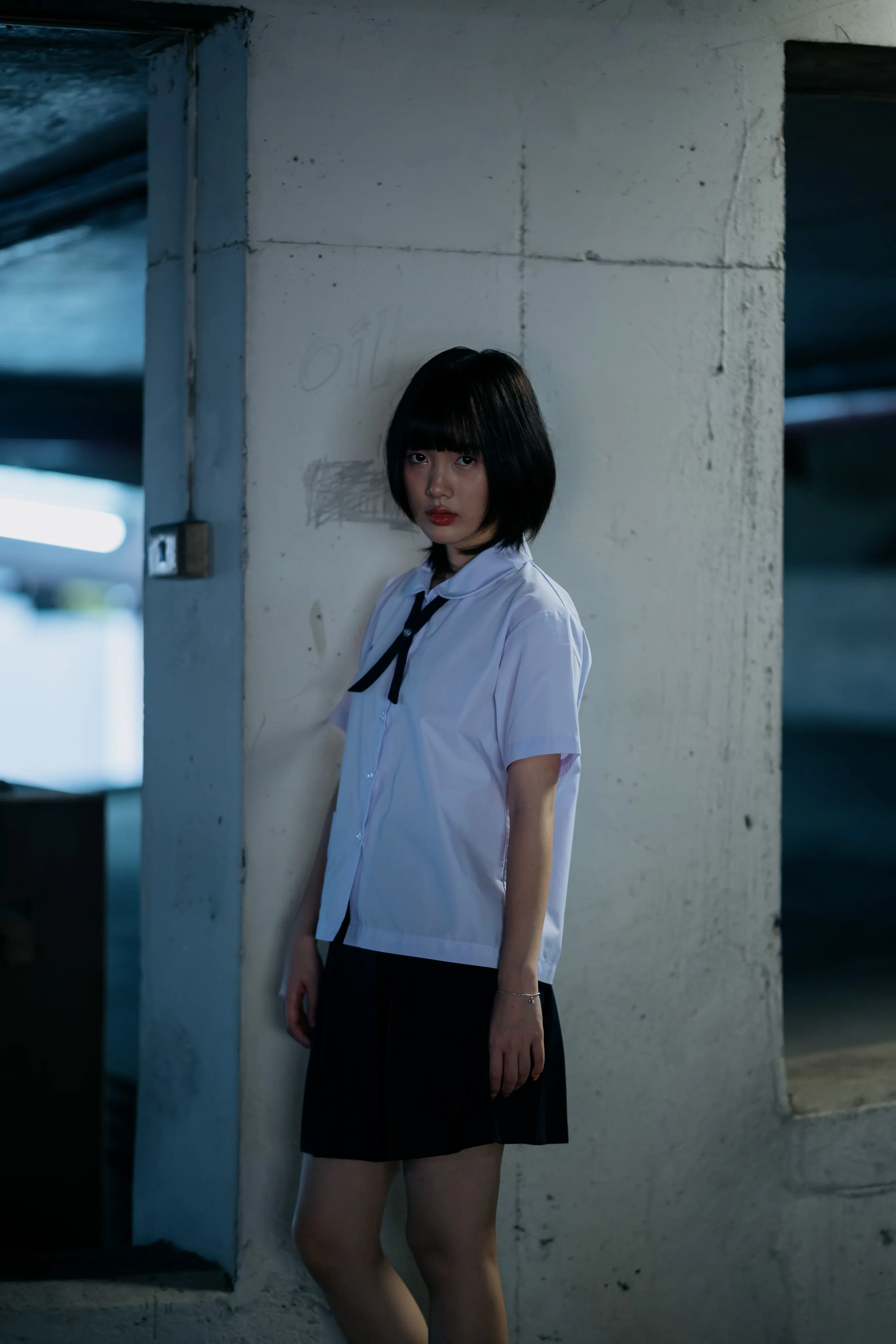 A girl in a school uniform leaning against a concrete pillar in a parking garage.