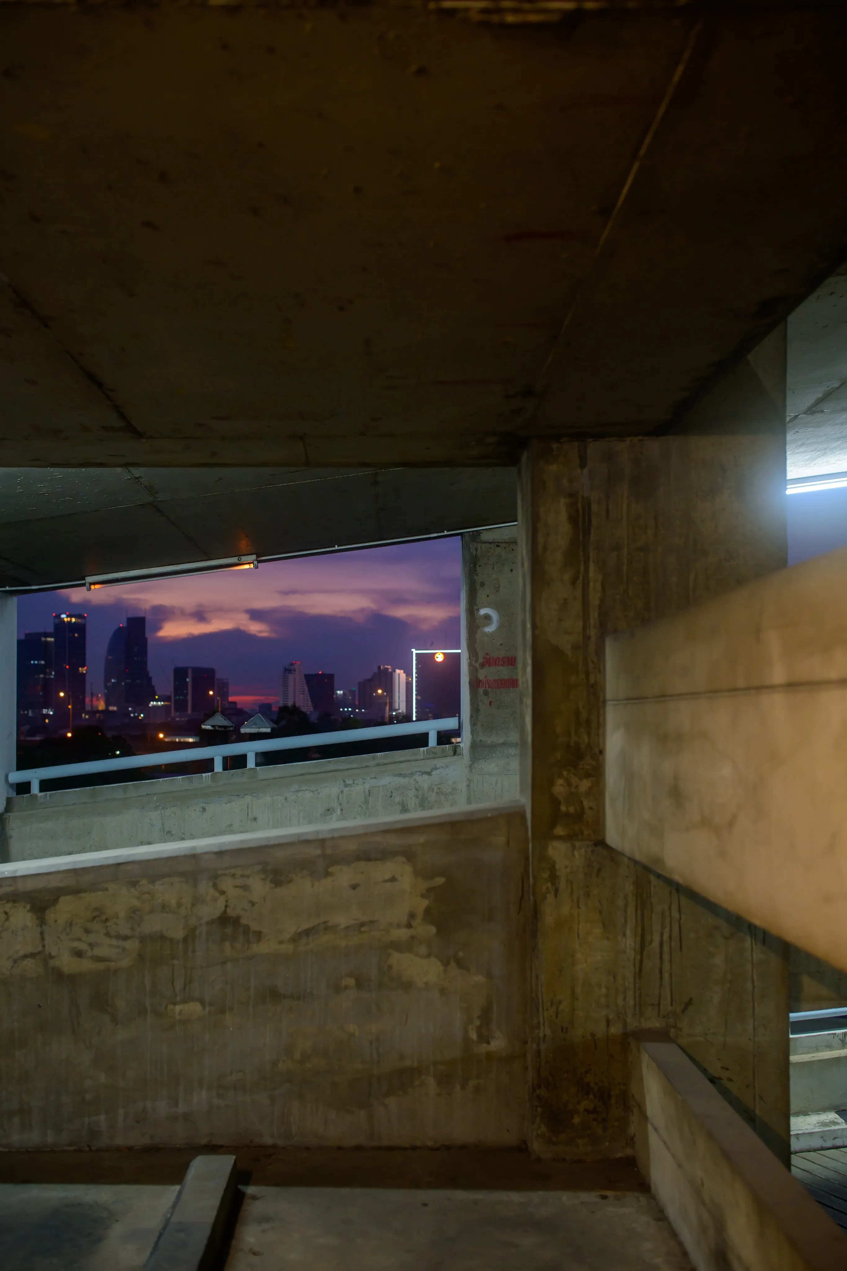 A view down a concrete parking garage stairwell toward a city skyline at dusk.