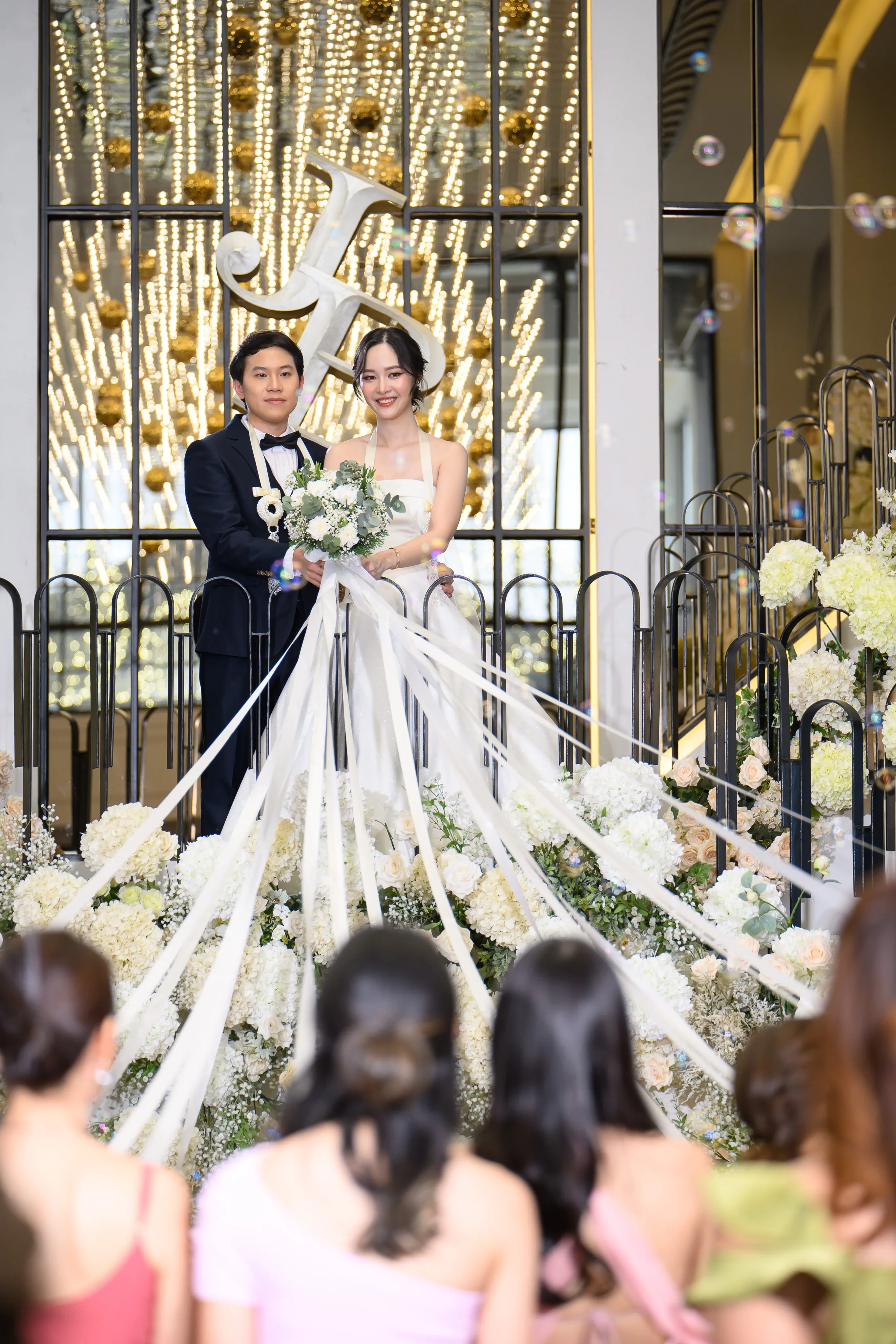 A bride and groom on a flower-lined stage as guests hold long white ribbons streaming from the bouquet