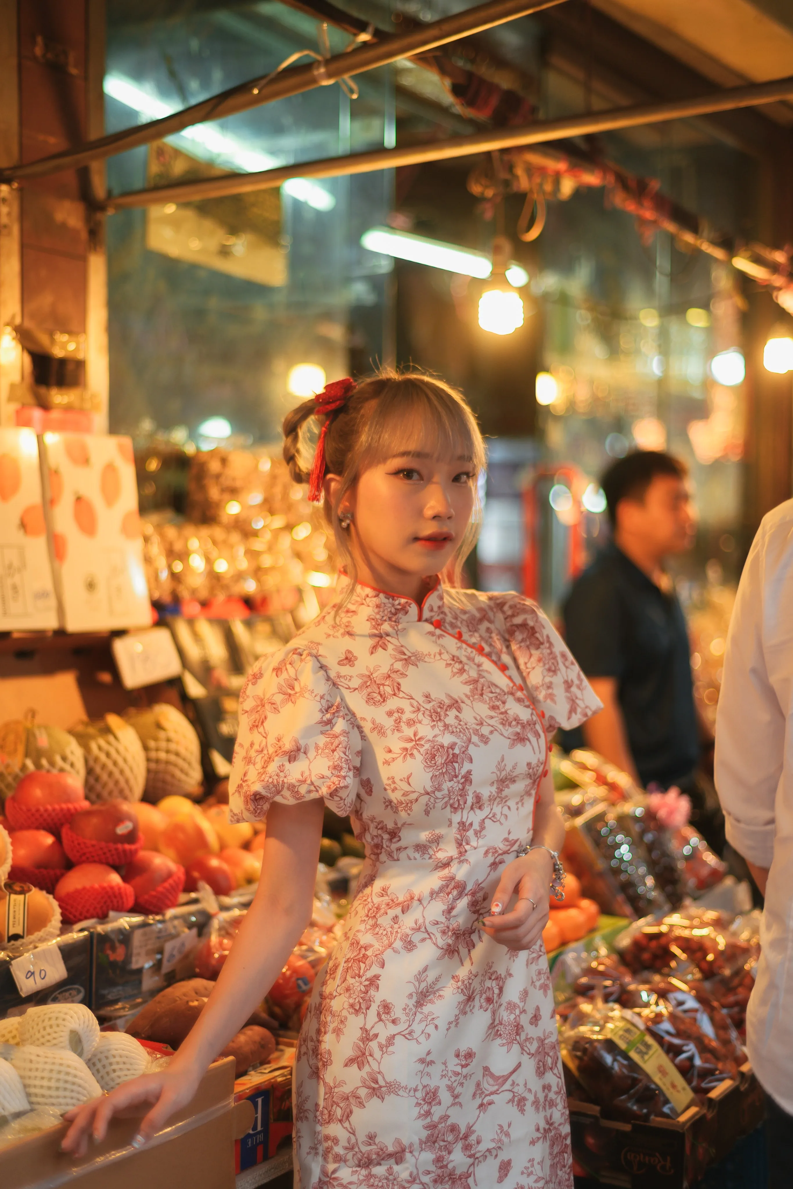 A woman in a floral cheongsam-style dress standing beside a lit fruit stall at a night market