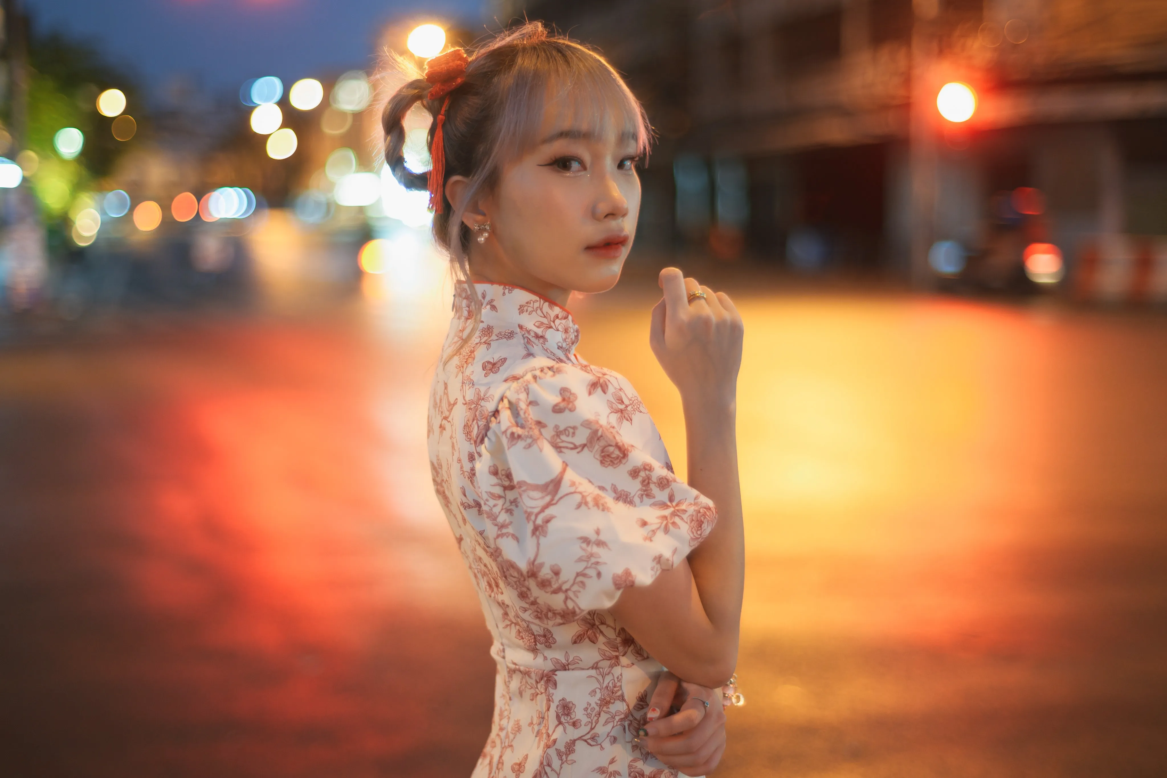 A woman in a floral cheongsam-style dress glancing over her shoulder against blurred city lights at dusk