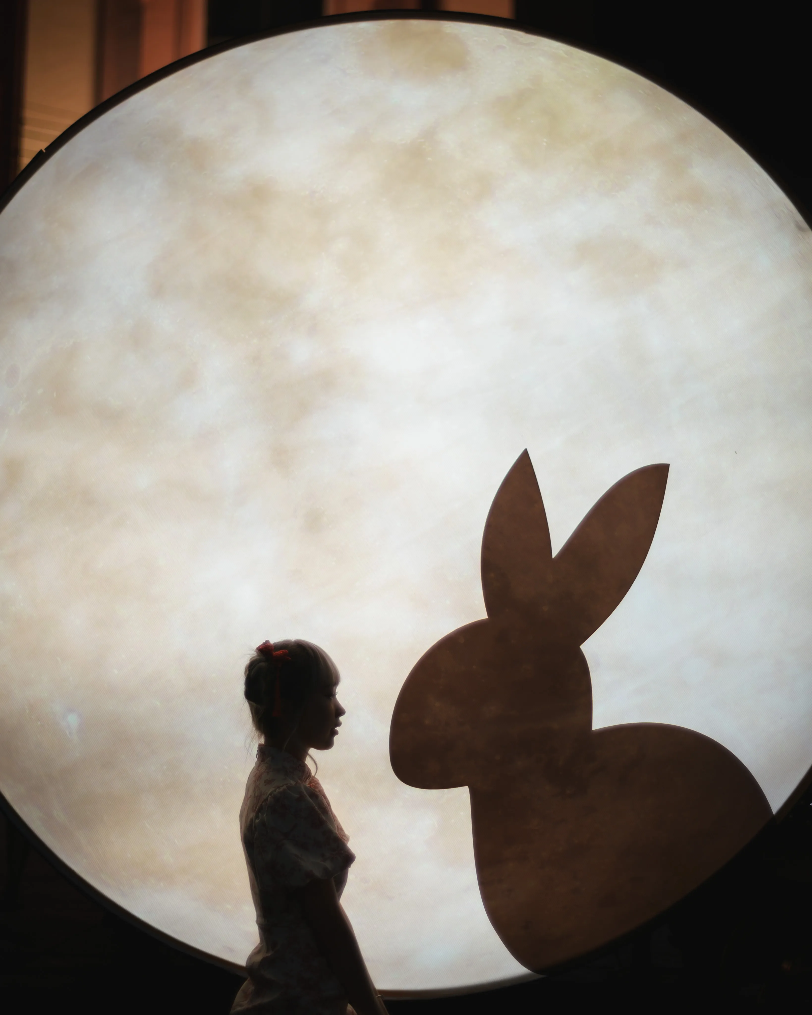 The silhouette of a woman beside a rabbit cut-out against a large glowing moon disc