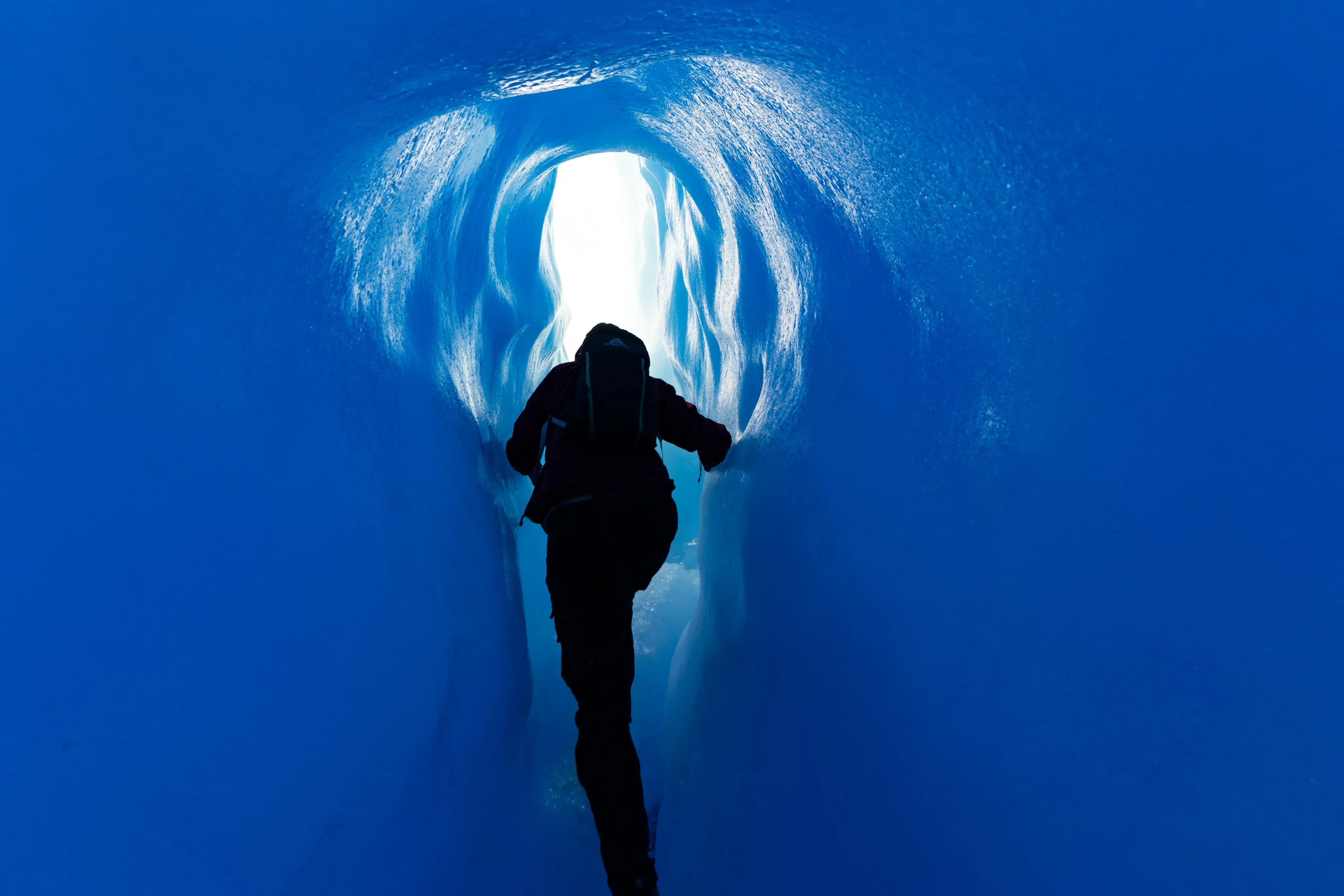 A silhouetted person climbing through a narrow blue ice tunnel toward a bright opening