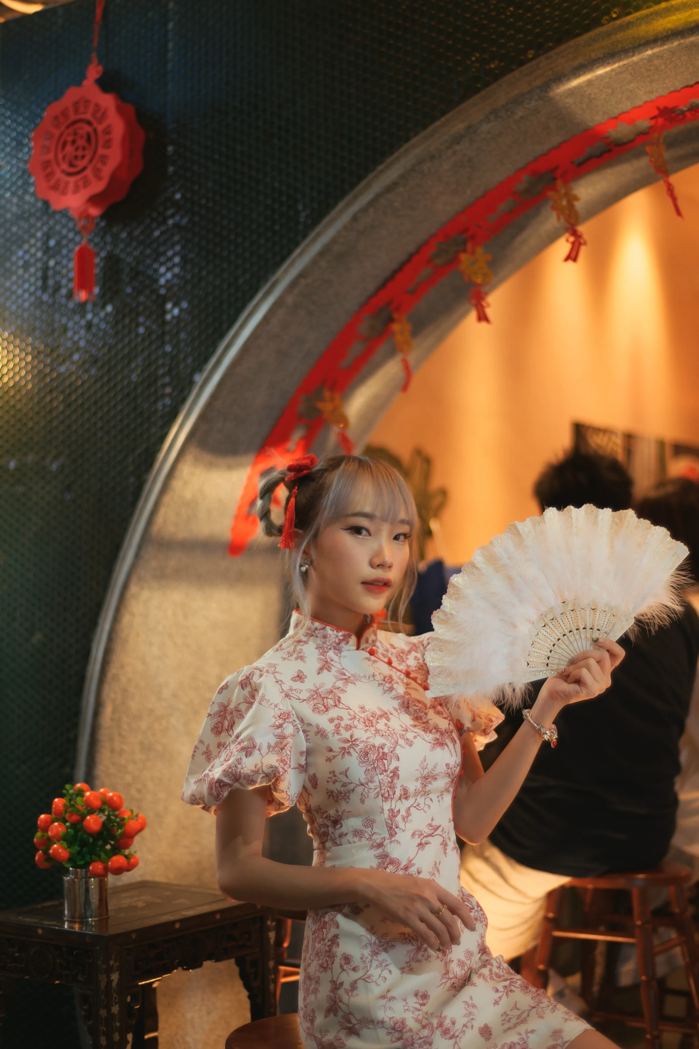 A woman in a floral cheongsam-style dress holding a white feather fan beneath a round red-trimmed archway