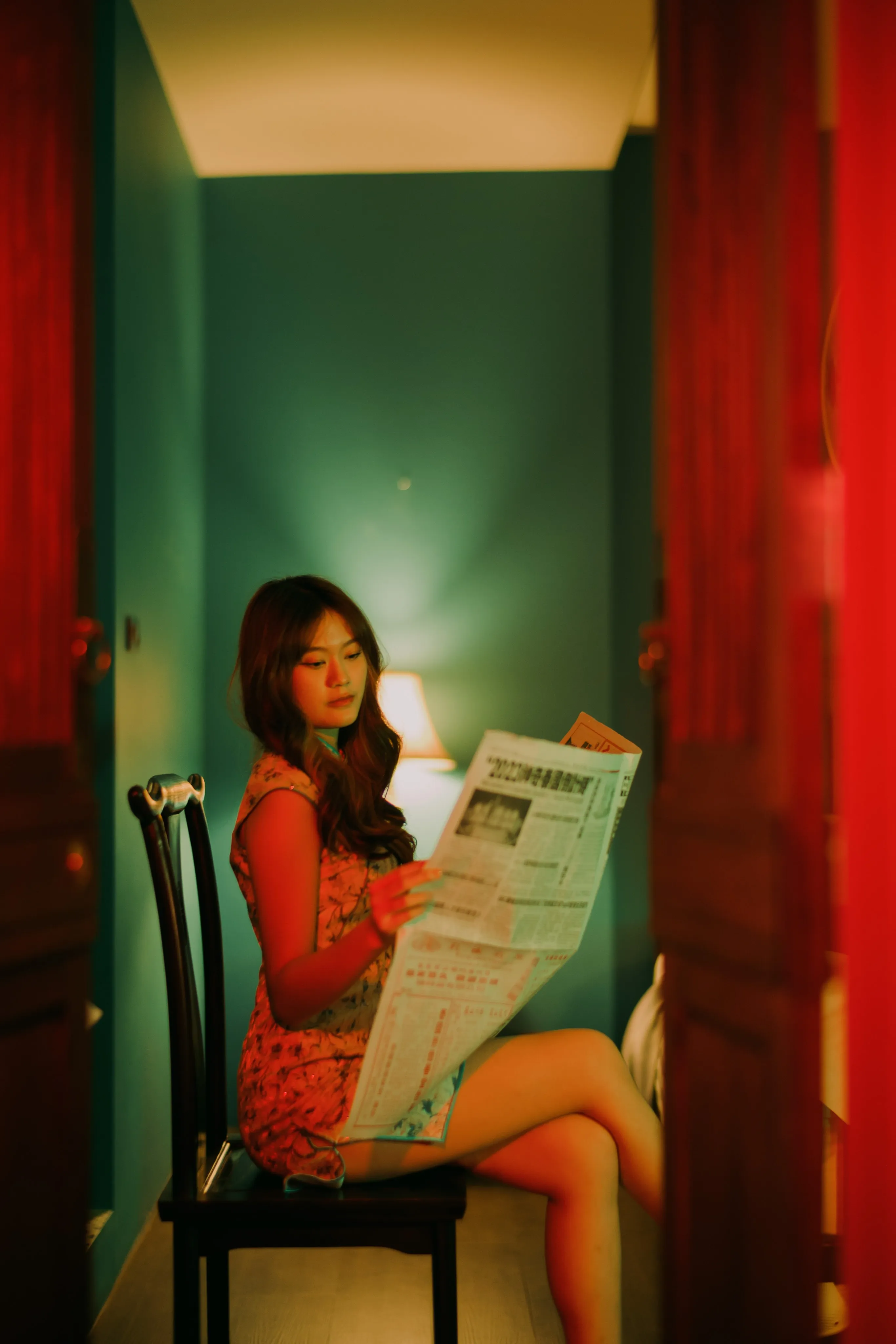 A woman in a floral qipao reading a newspaper in a chair, framed by red doors in a green-walled room