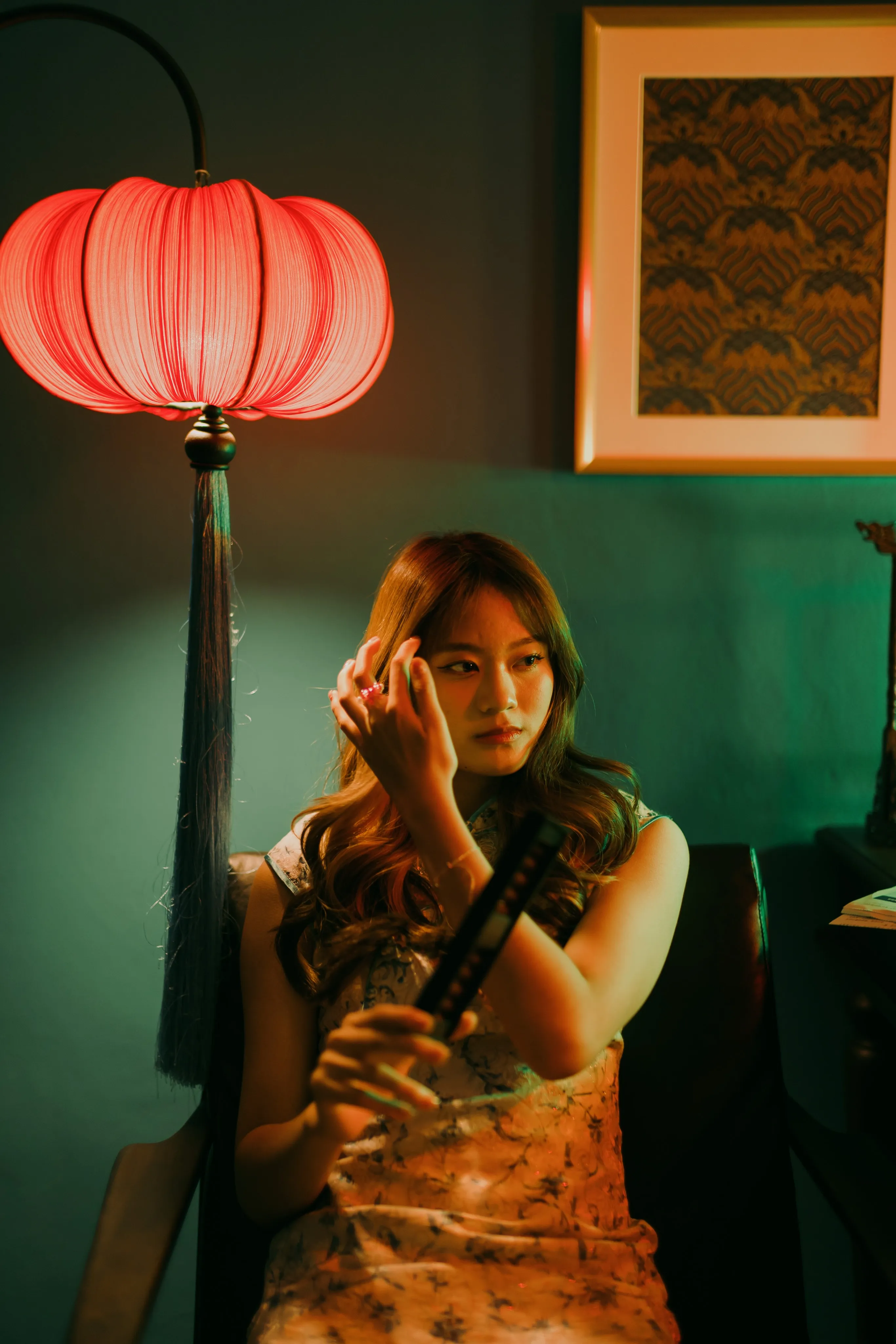 A woman in a qipao holding a folding fan beside a glowing red lantern lamp in a green room