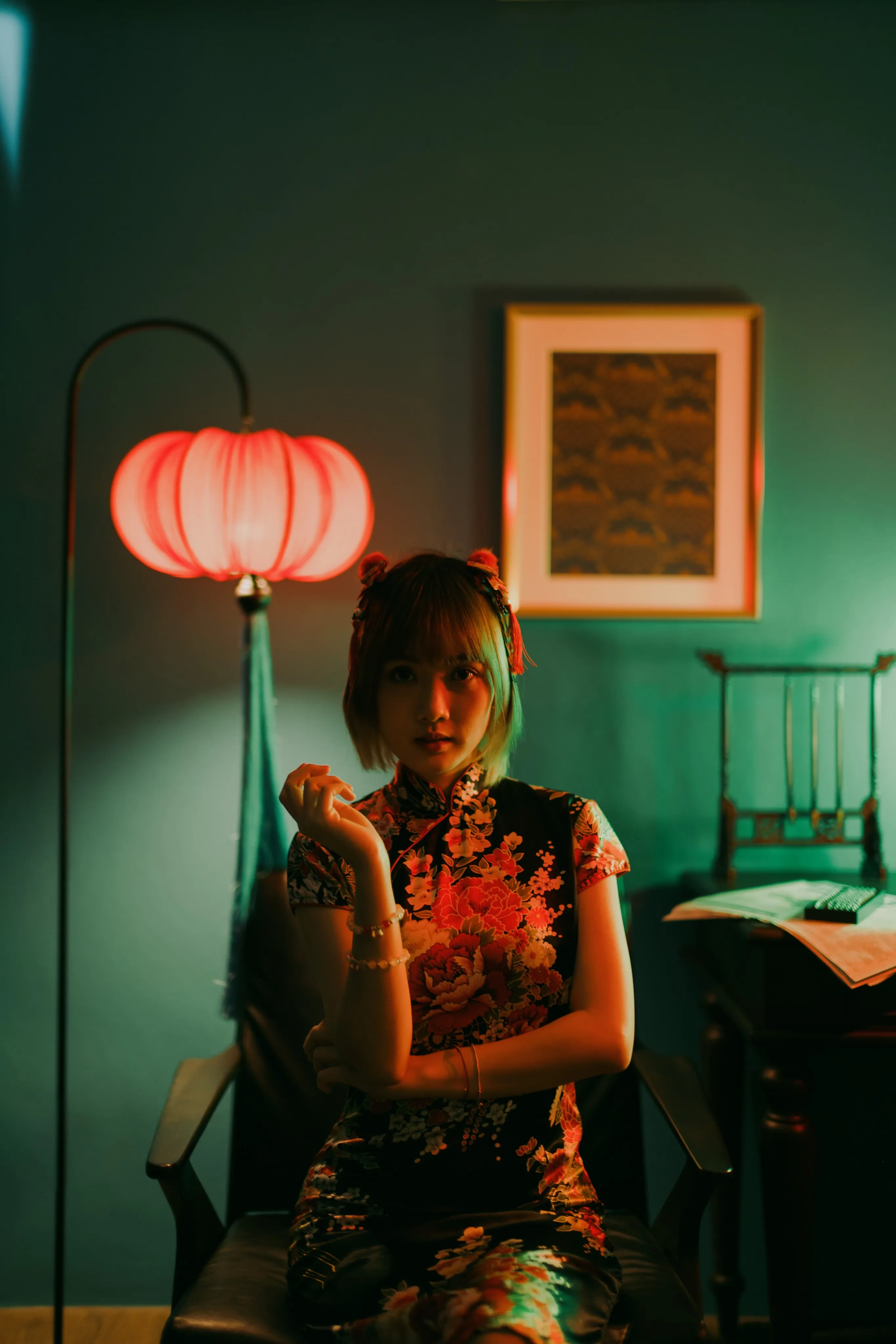 A woman in a black floral qipao seated beside a red lantern floor lamp against a teal wall